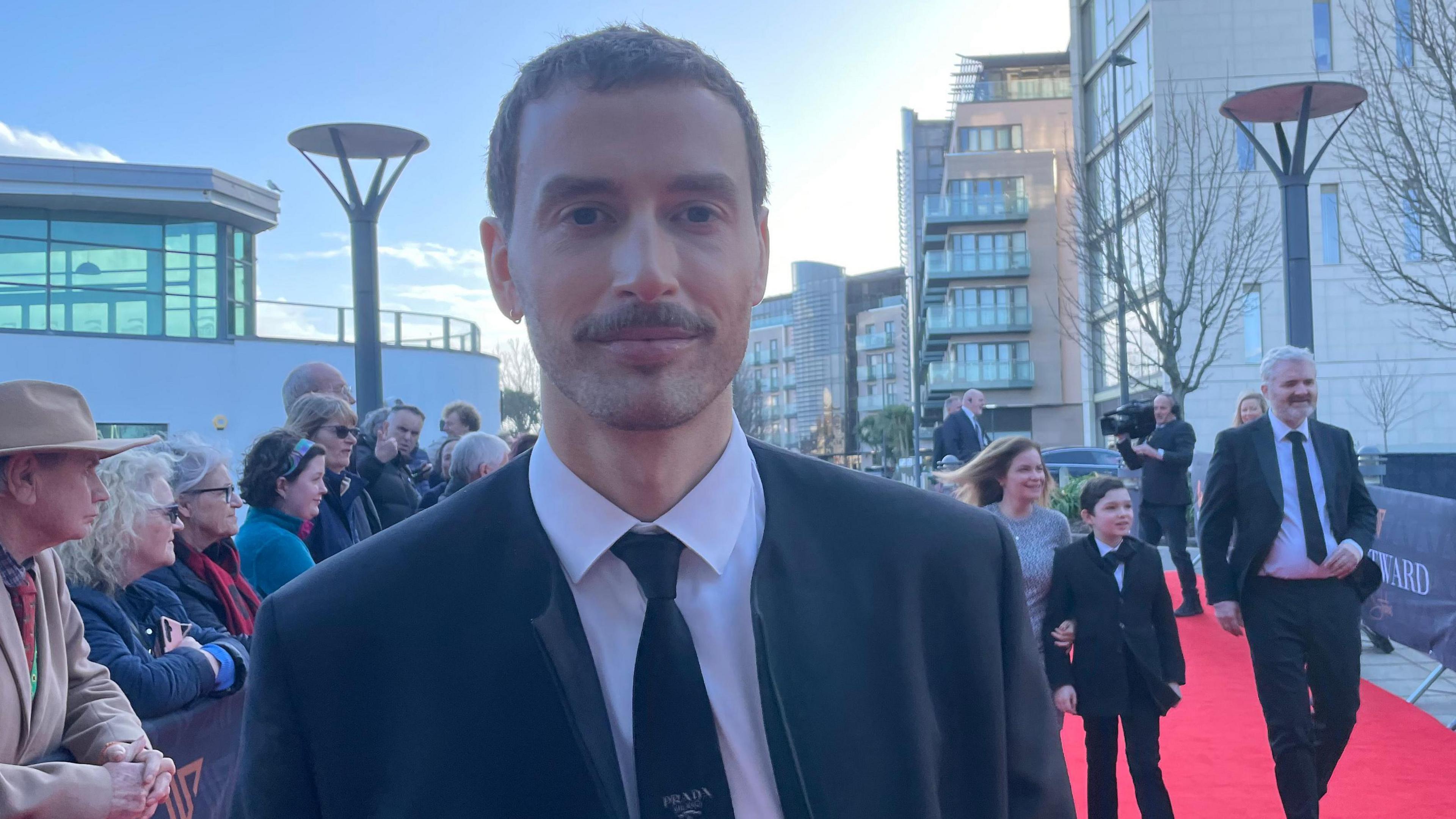 Timothy Renouf is standing on the red carpet smiling at the camera. He is wearing a white shirt with a black tie and a black collarless jacket. He has short brown hair, a moustache and a slight beard. In the background a woman, man and child can be seen walking along the red carpet. There are spectators leaning on a barrier behind the actor.