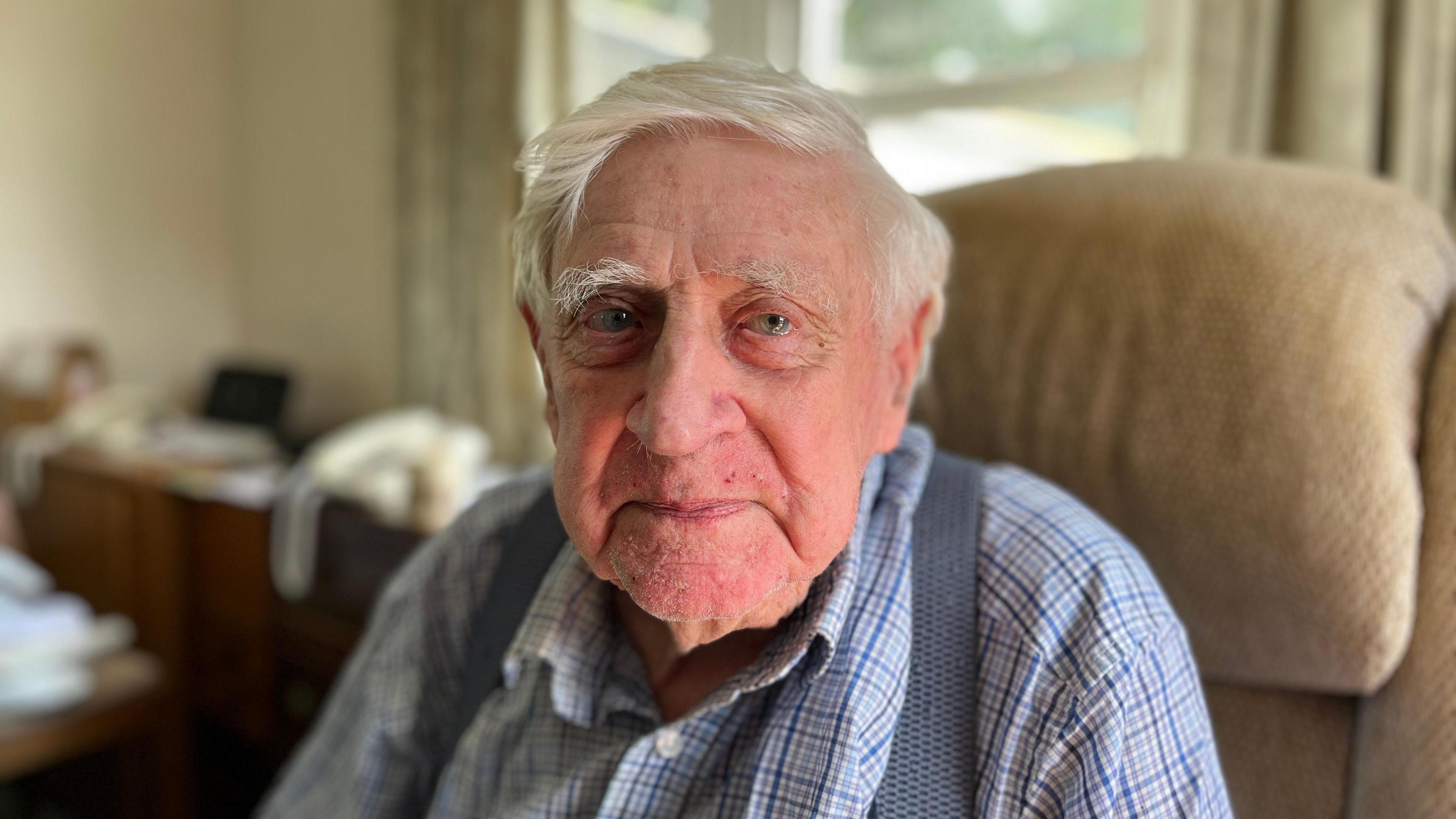 Wilfred John Brett wearing an unbuttoned chequered shirt and braces sitting in an arm chair looking at the camera. He has grey hair and behind him are two landline phones