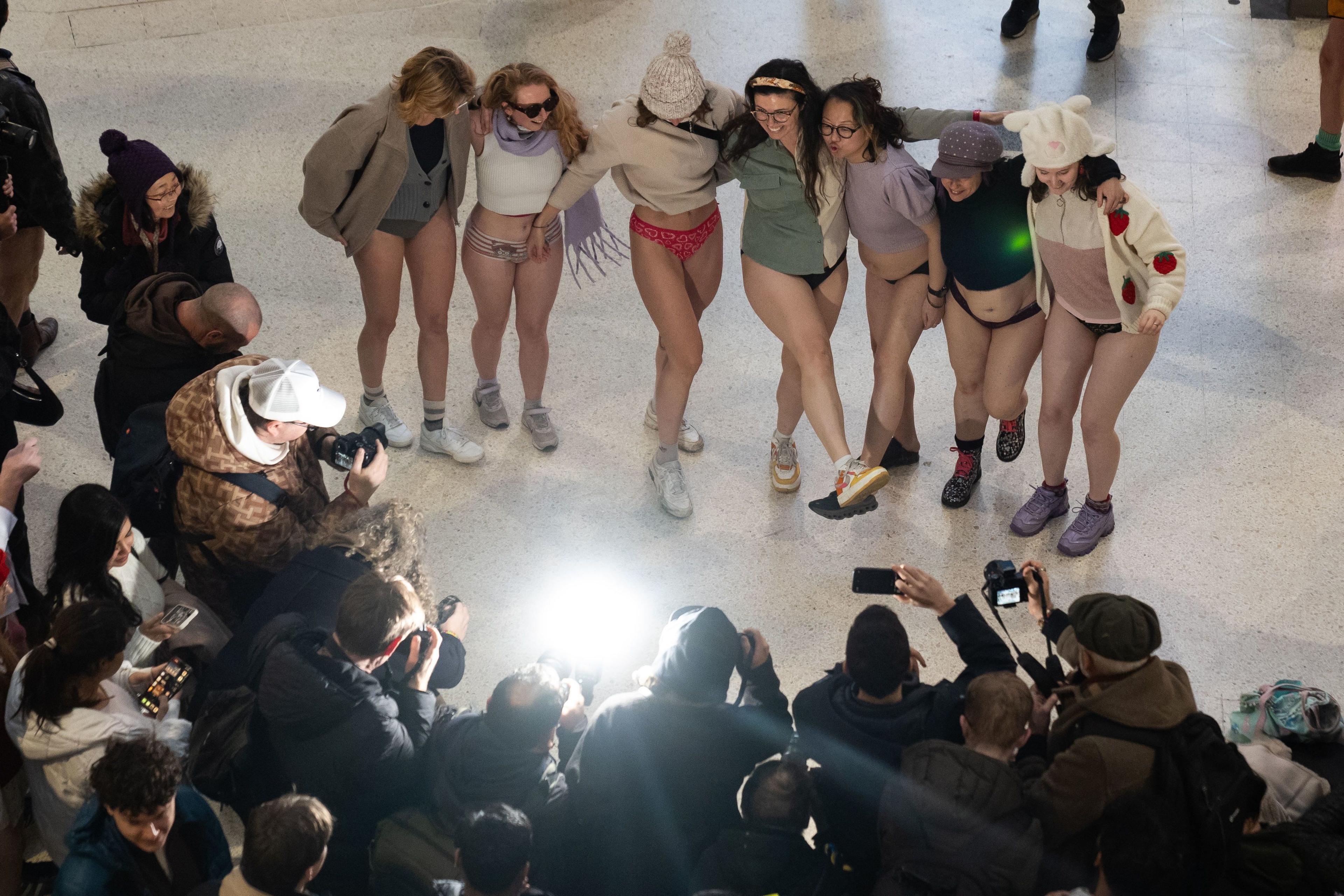 Seven women not wearing trousers with arms around each other stand on the concourse at Waterloo station while photographers and members of the public for a semi-circle to take pictures