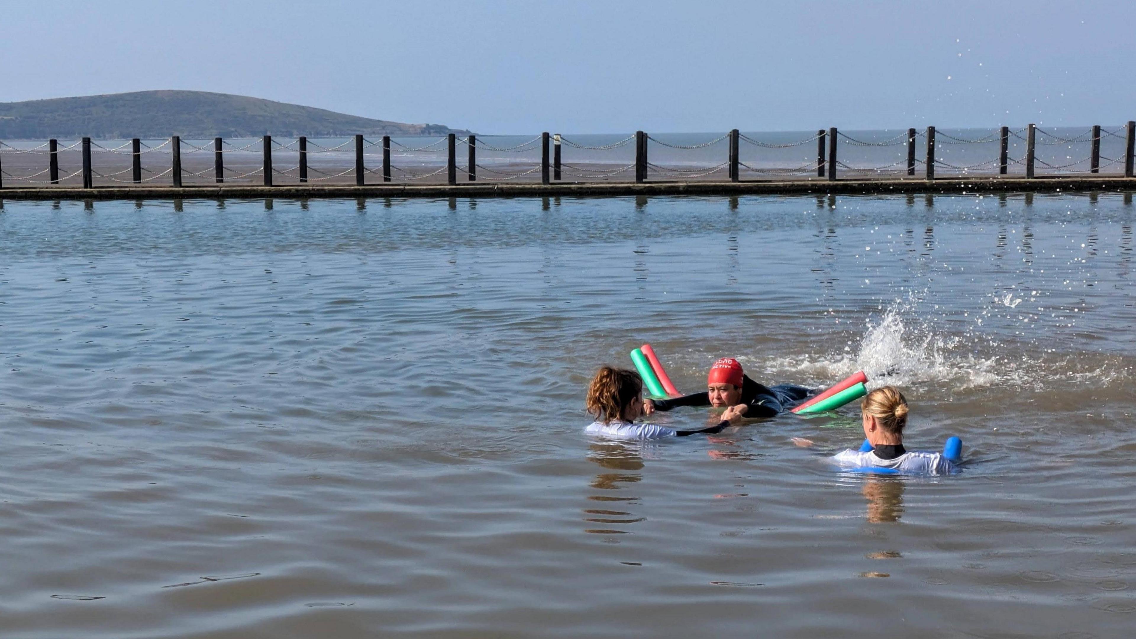 A woman wearing a black wetsuit and red swimming cap. She is lying on her front on top of two pool noodles to help her float in Weston-super-Mare Marine Lake. She is kicking her legs and there is a lot of water splashing behind her. Two women wearing white rash vests are in front of her, with one pulling her arms to help her go forward.