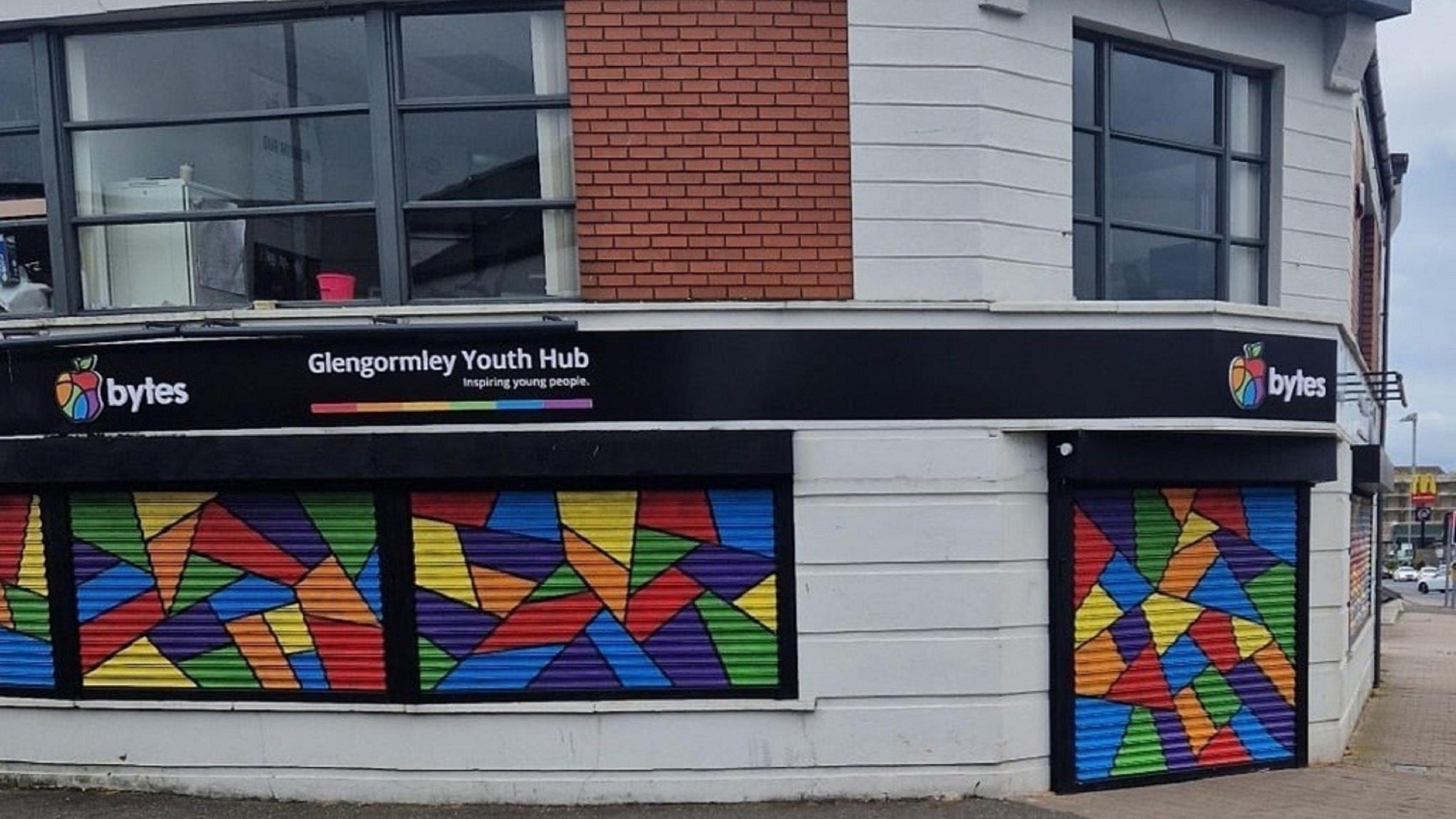 A building on a corner made of red bricks and white concrete blocks. There are large windows on the first floor. The ground floor windows and main entrance are covered with multi-coloured shutters