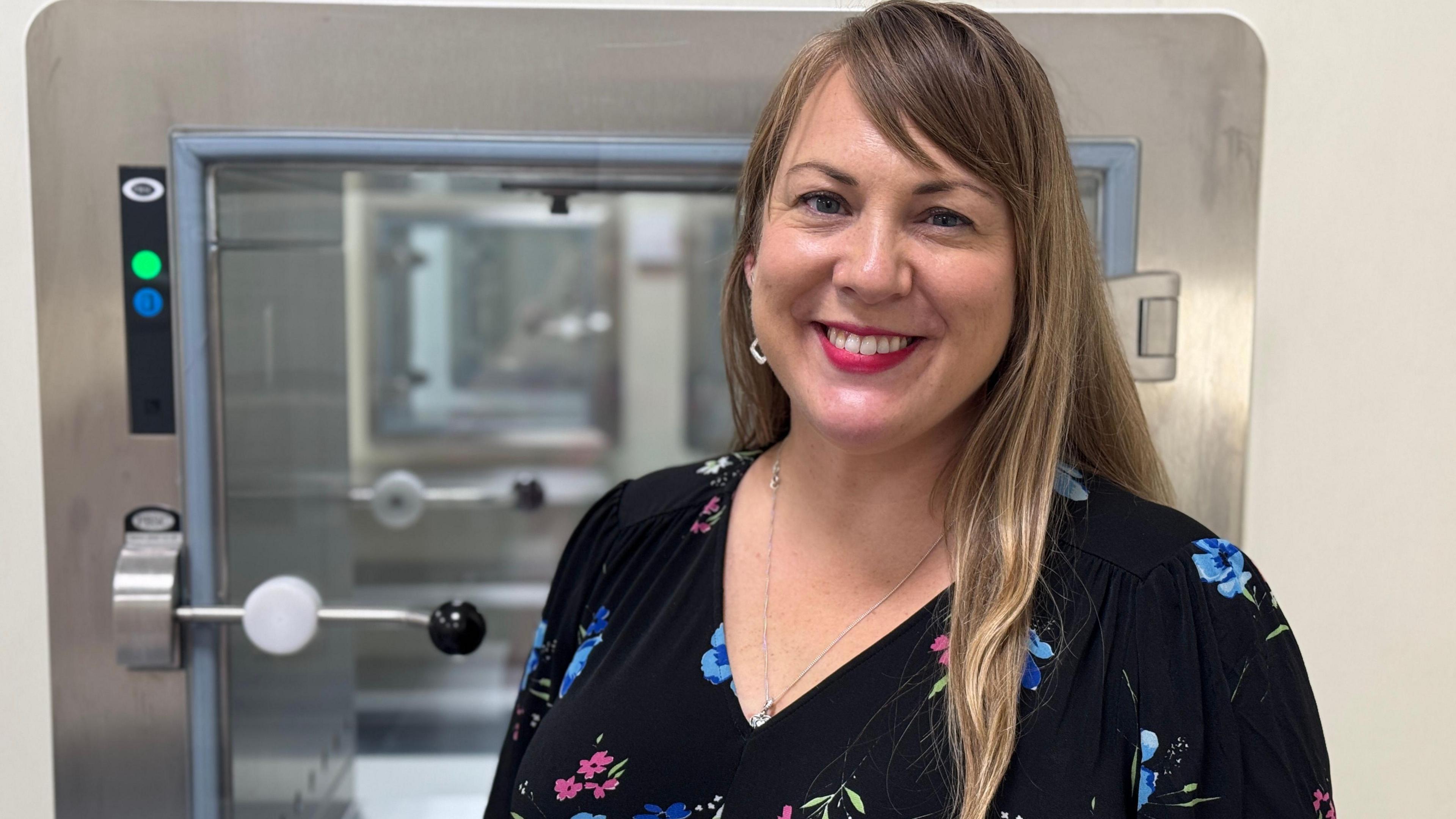 The lab's director, Sarah Albon. She has long blonde highlighted hair and is wearing pink lipstick and a black floral dress. She is smiling into the camera. Behind her is a piece of equipment.