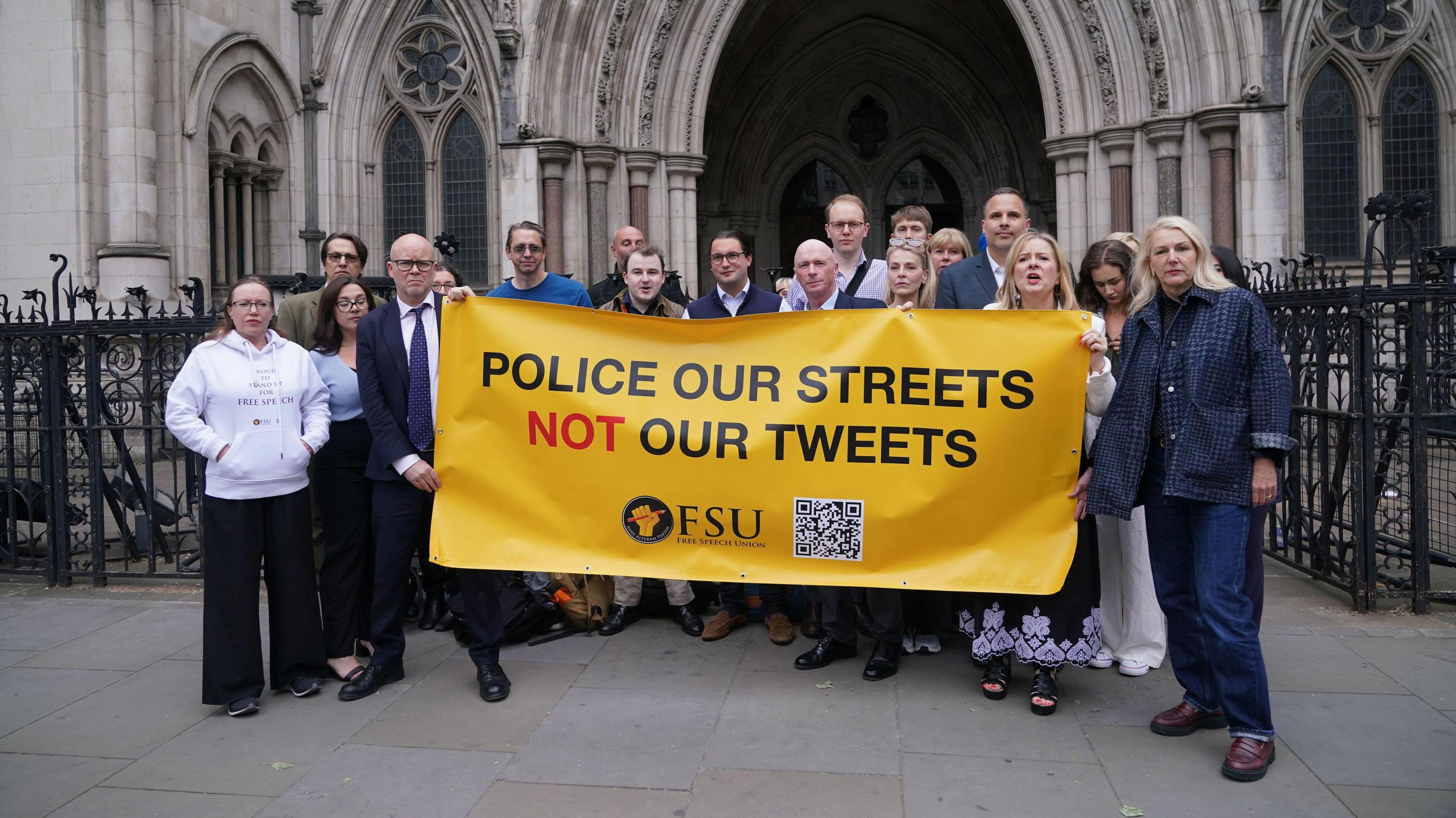 A group of people standing outside the Royal Courts of Justice holding a yellow banner with the slogan "police our streets not our tweets" written in capital letters. The words are mostly black expect for "not", which is in red. The banner includes the Free Speech Union logo, which is a fist clutching a sharpened pencil and the letters FSU. There is also a black and white QR code on the banner.