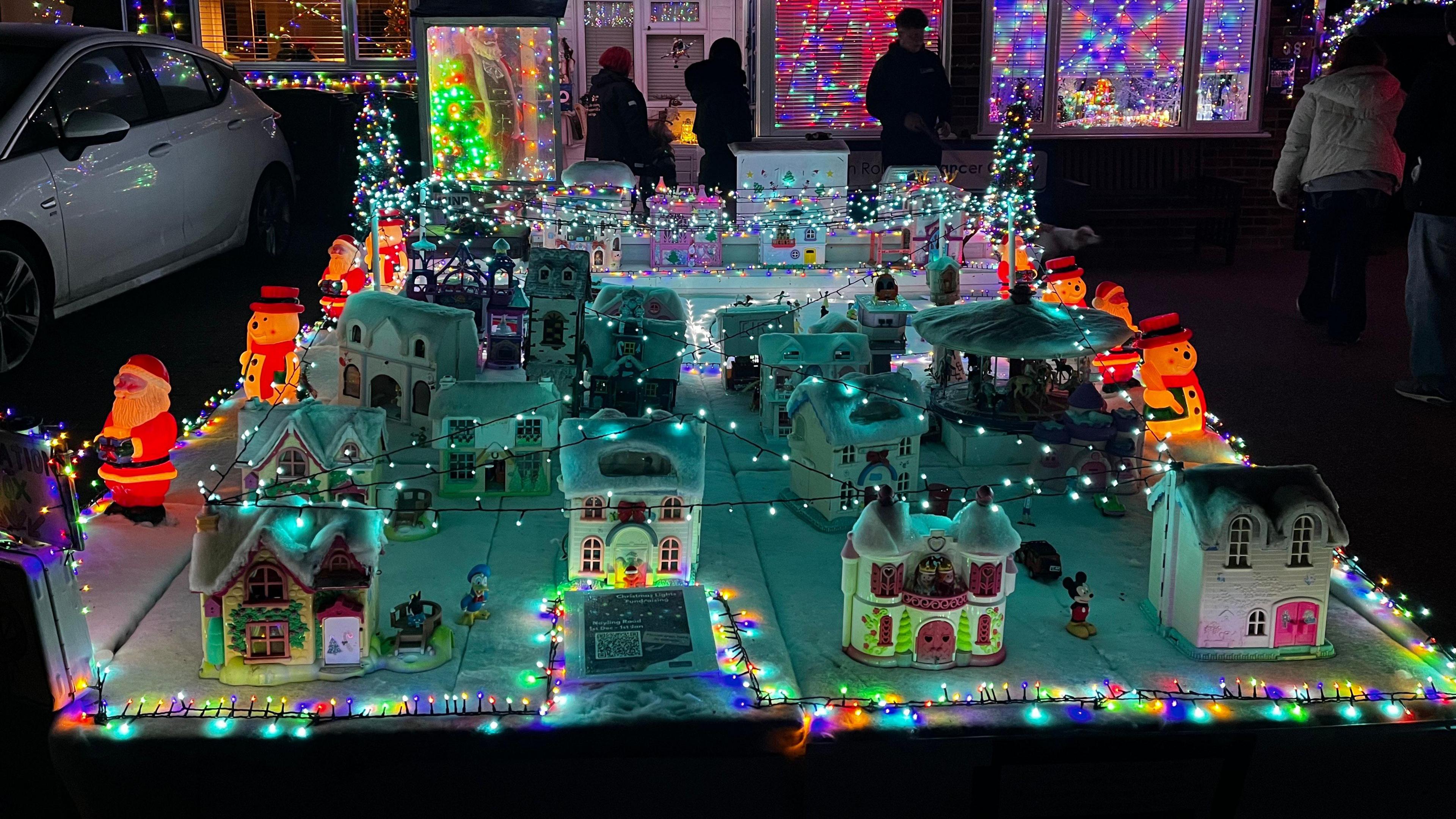 About 13 miniature houses are on display on a large square blanket of fake snow, adorned with fairy lights and Disney figurines. Illuminated snowmen and Father Christmas figures are lined up along each side. People are standing and looking at the display in the background.