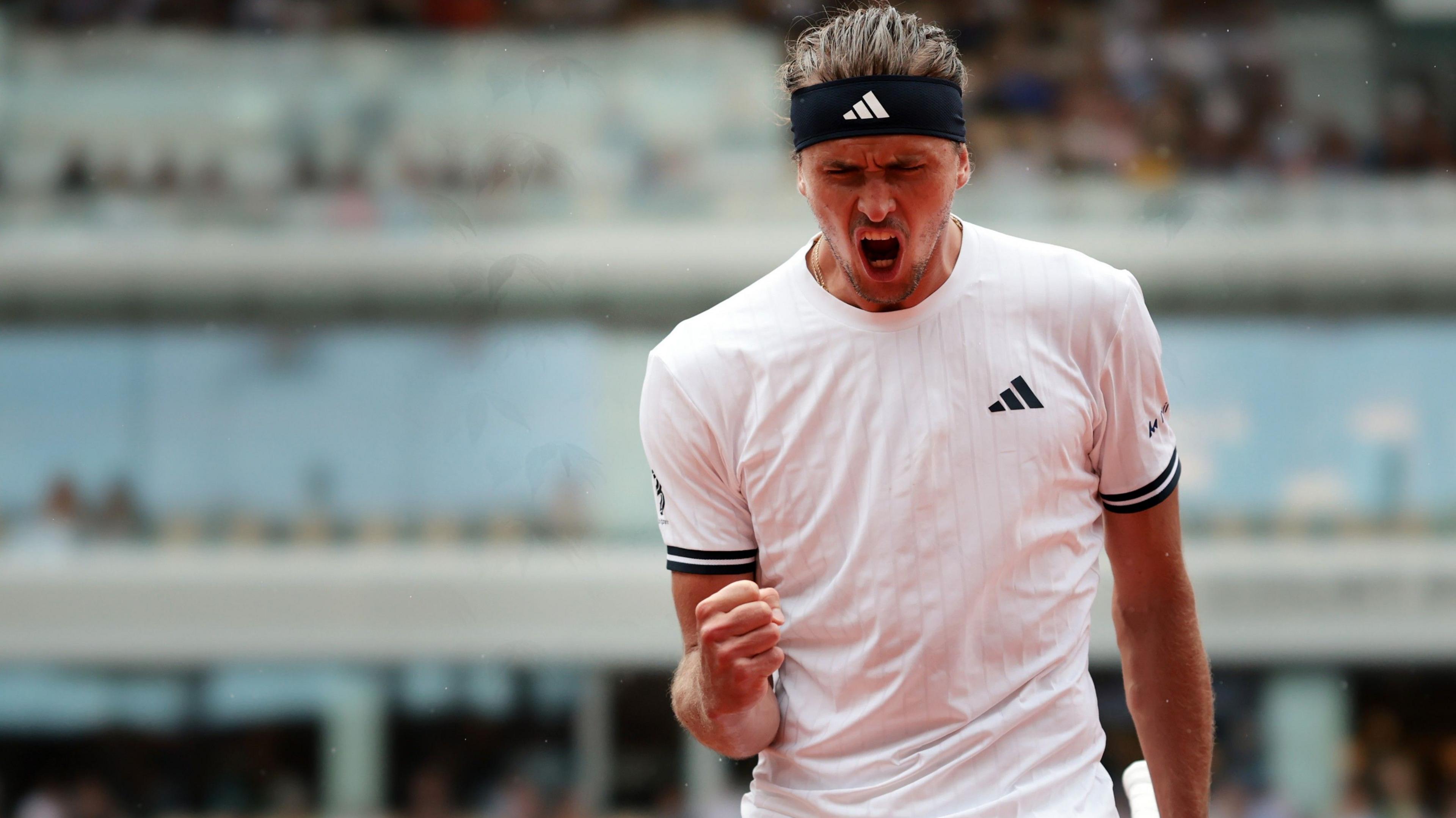 Alexander Zverev, wearing a white t-shirt and black headband, roars with his mouth wide open and clenches his right fist in celebration