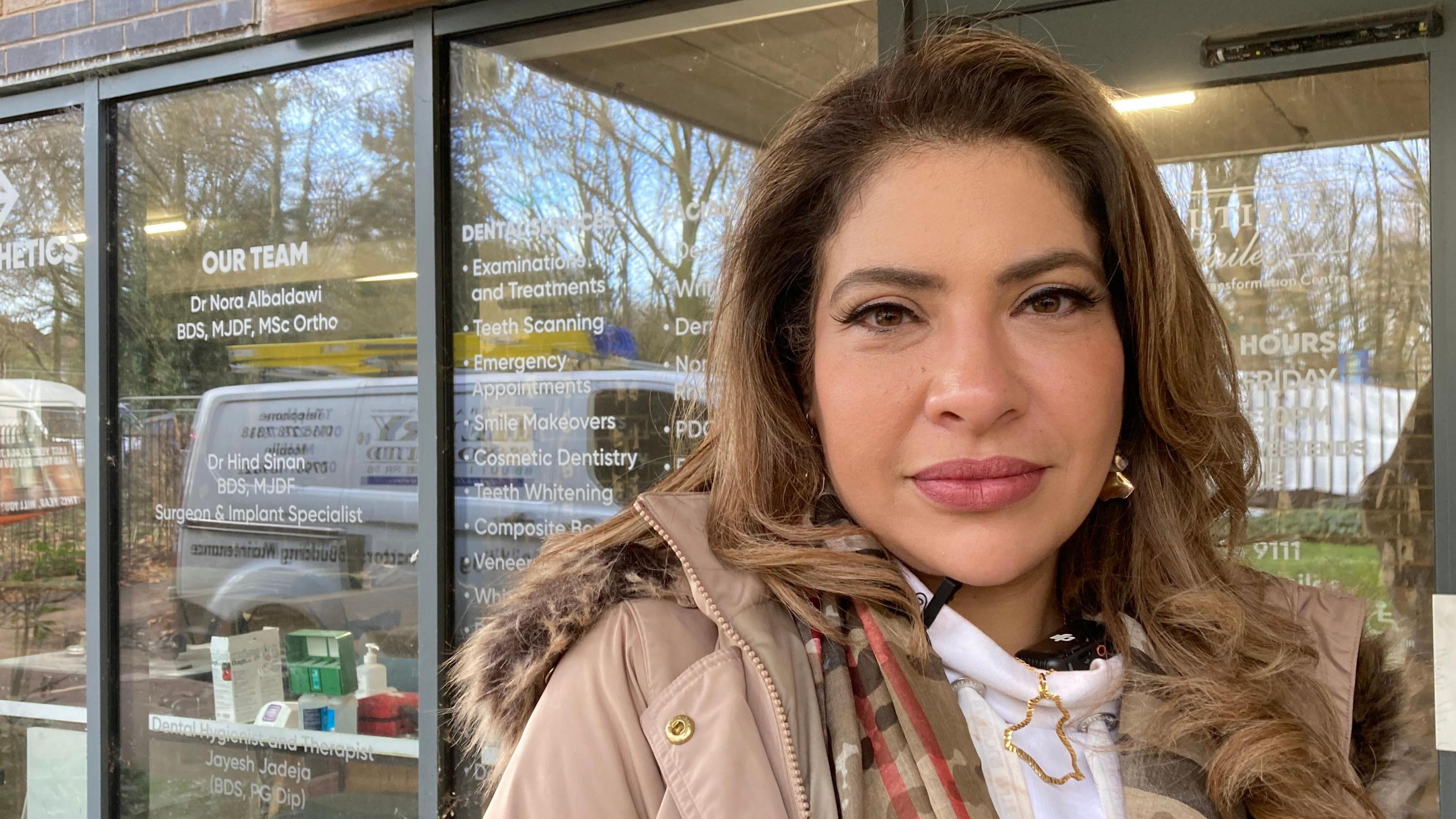 With Brown hair and wearing earrings, Dr Nora, wearing a brown jacket and multi-coloured scarf, looks at the camera as she stands in front of her new dental practice