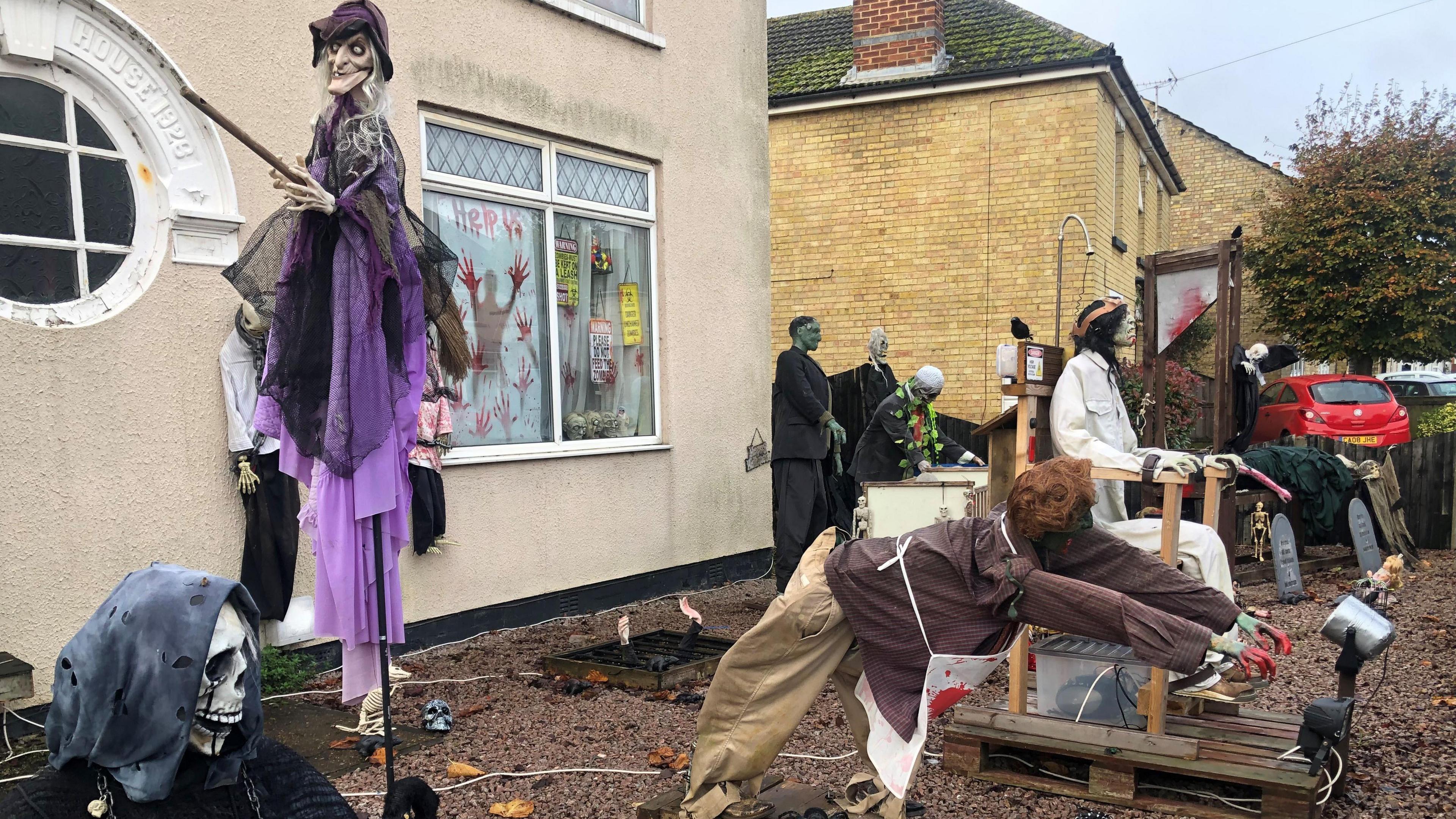 A witch suspended as though in flight on a broom stick in a gravelled front garden with an assortment of life-size figures including Frankenstein's monster, a guillotine, an electric chair, a hooded skeleton is sitting on the left and there are some gravestones on the right. The two-storey house behind is cream coloured with a  panelled round window on the left and a rectangular window on the right.