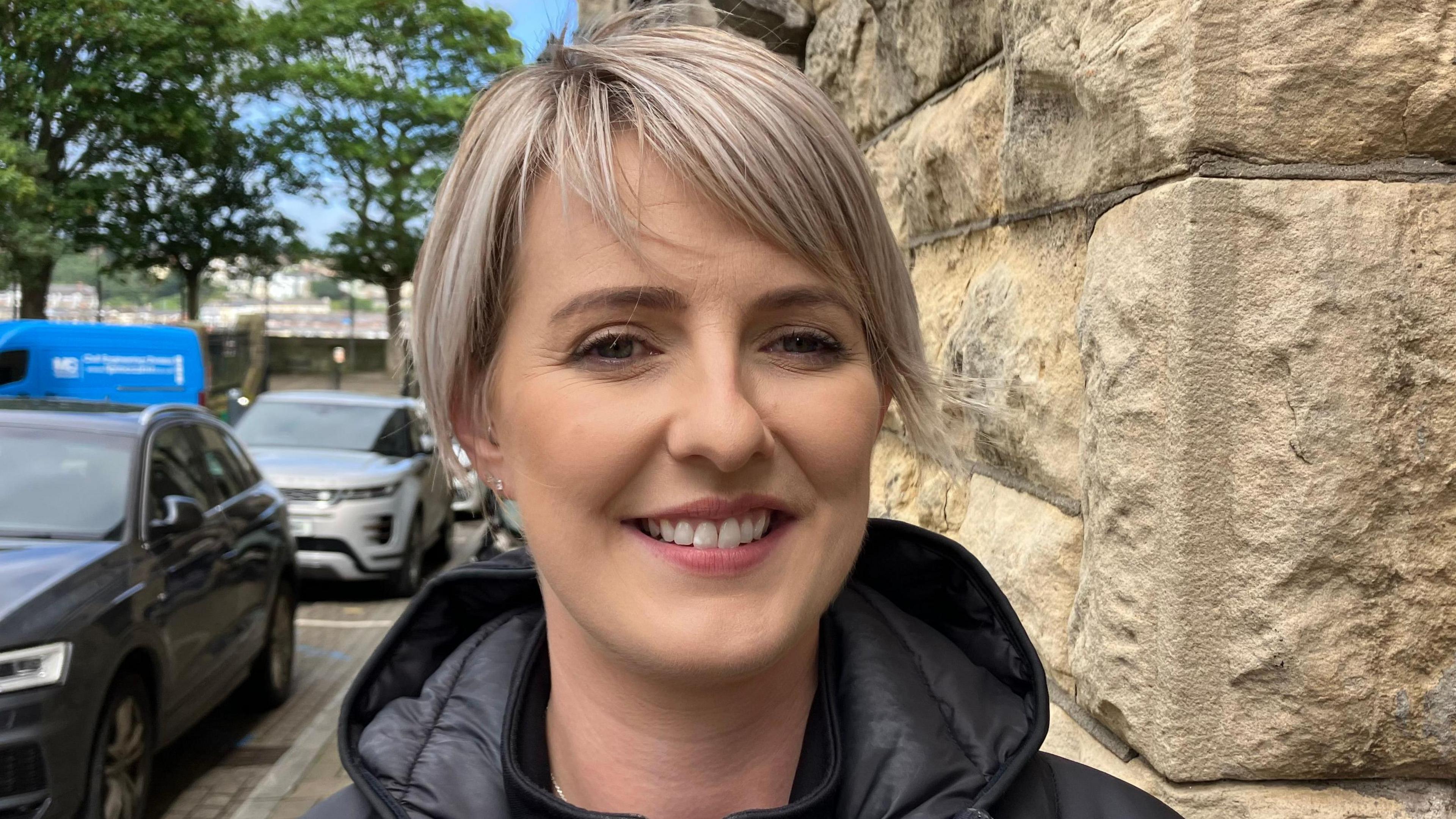 Georgina Kee-McCarter has short blonde hair and is wearing a black hooded coat. She is standing beside Derry's historic walls and cars parked on the street.