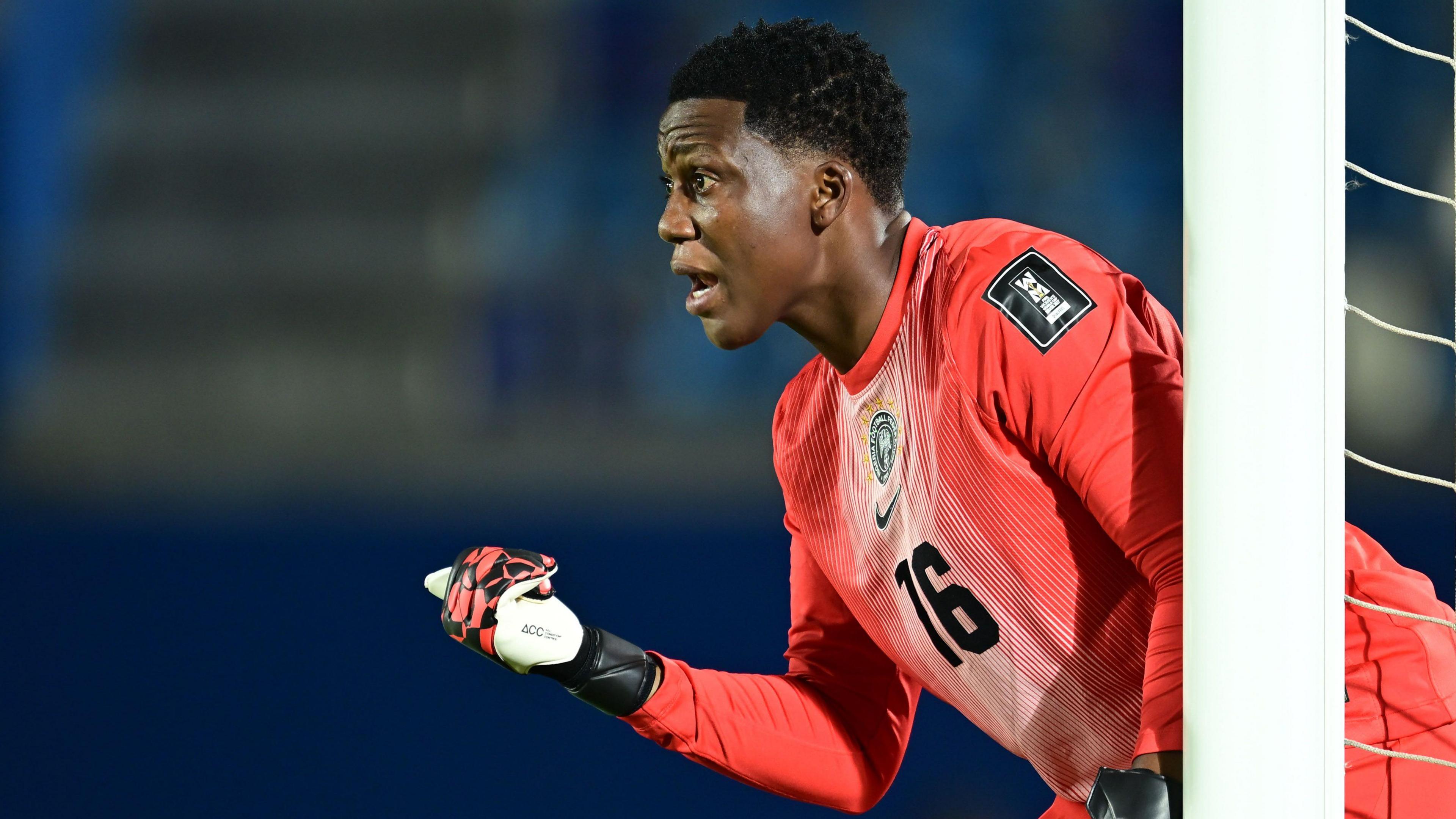 Chiamaka Nnadozie, wearing goalkeeping gloves and a bright orange shirt with a black number 16 on the chest, points her right thumb in front of her as she looks to arrange a defence which is out of picture. She stands by a white goal post, with netting visible at the side of the shot