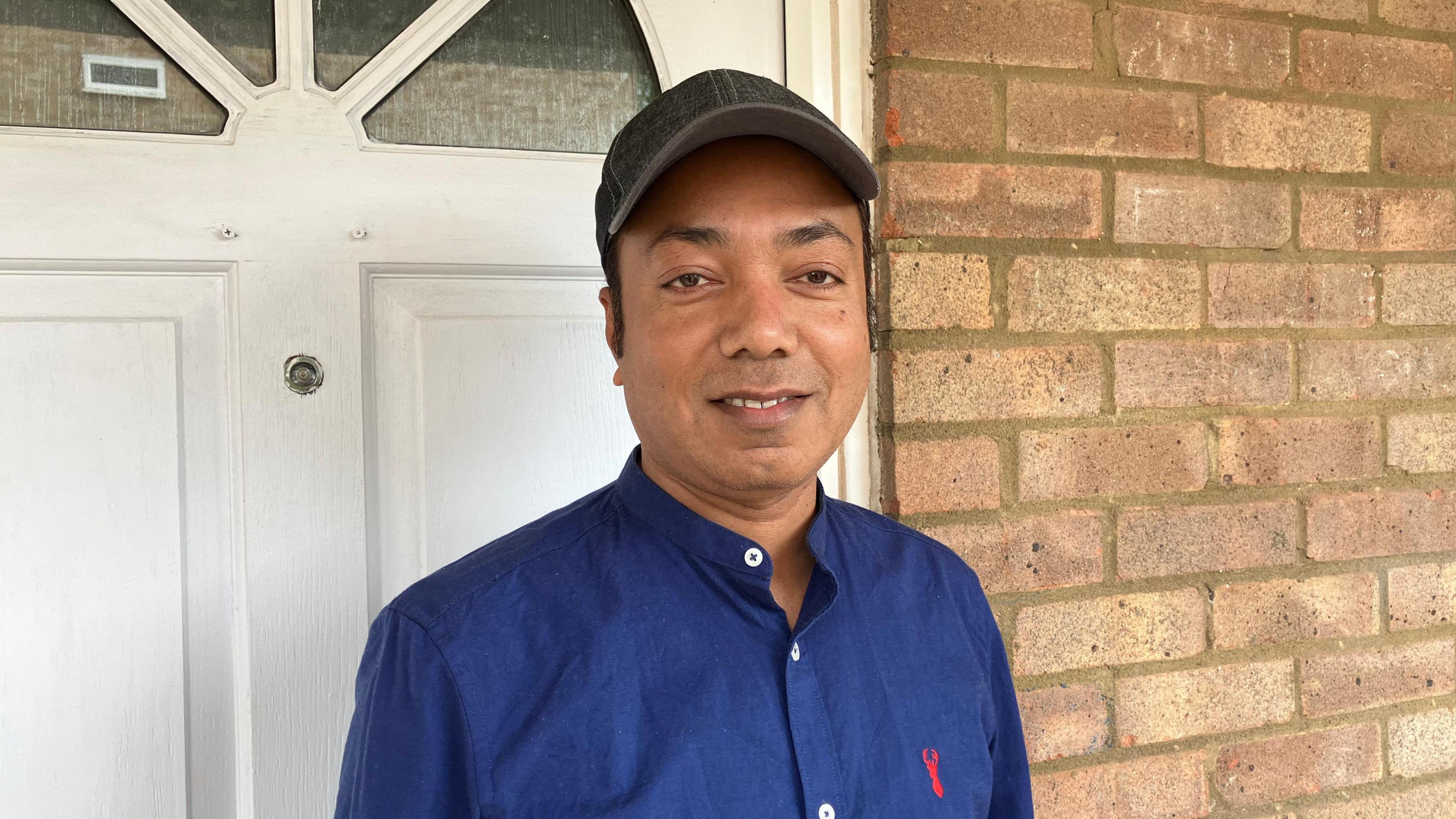 Zalal Abdul in a blue shirt standing in front of a white door and a brick wall