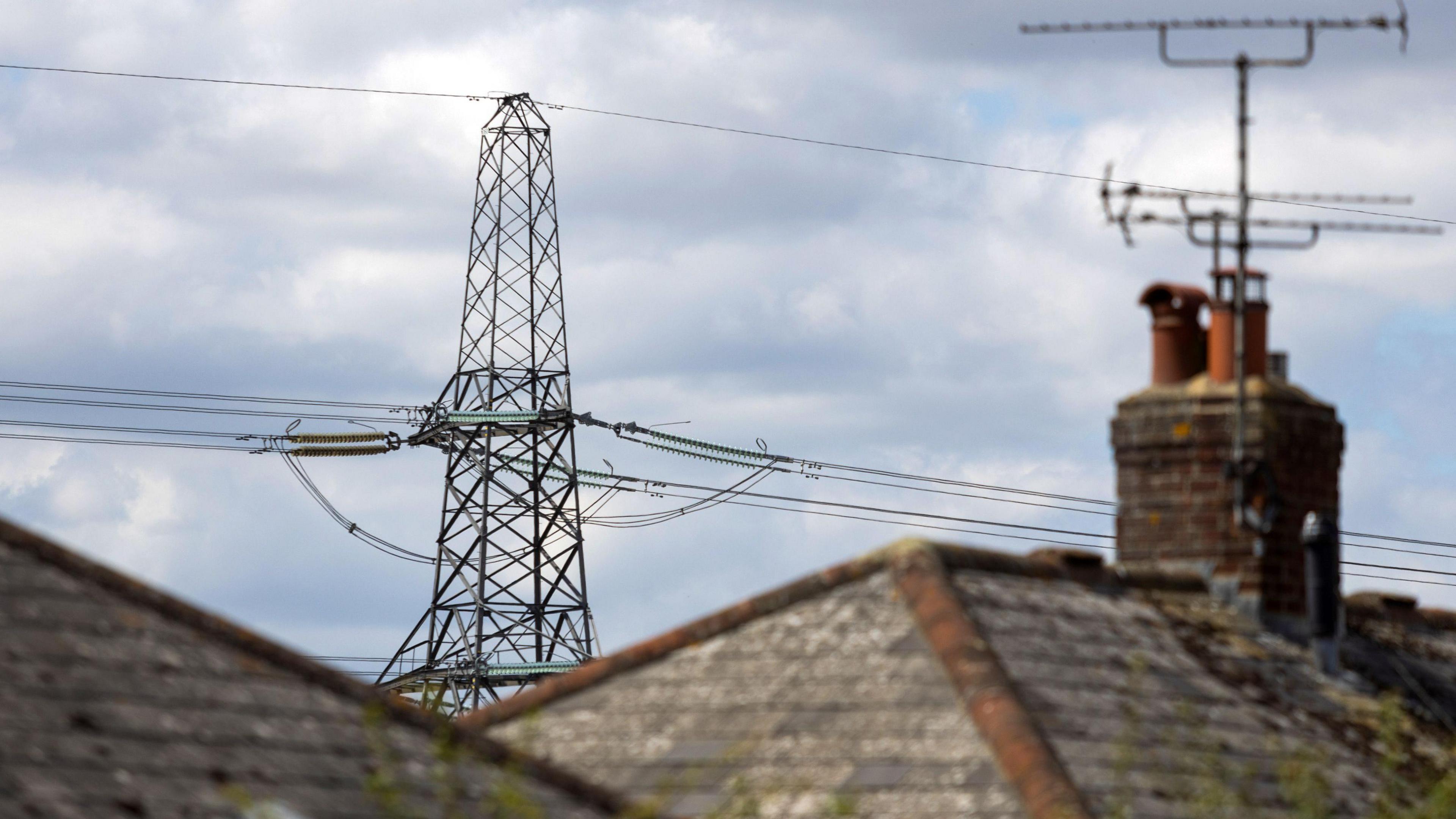 An electricity transmission tower and behind rooftops under a cloudy sky.