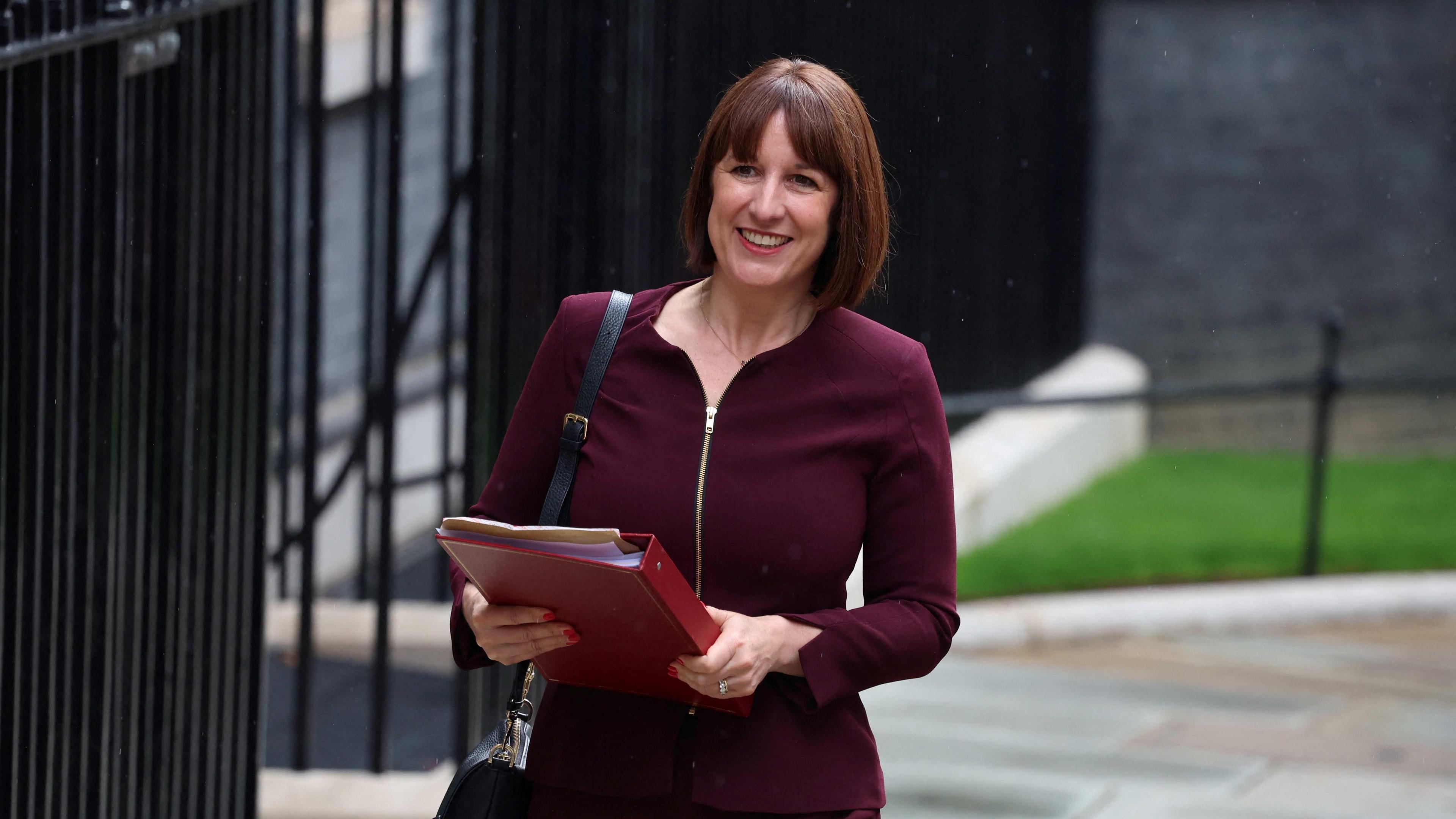 Rachel Reeves wearing a burgundy jacket and skirt is pictured carrying a red folder