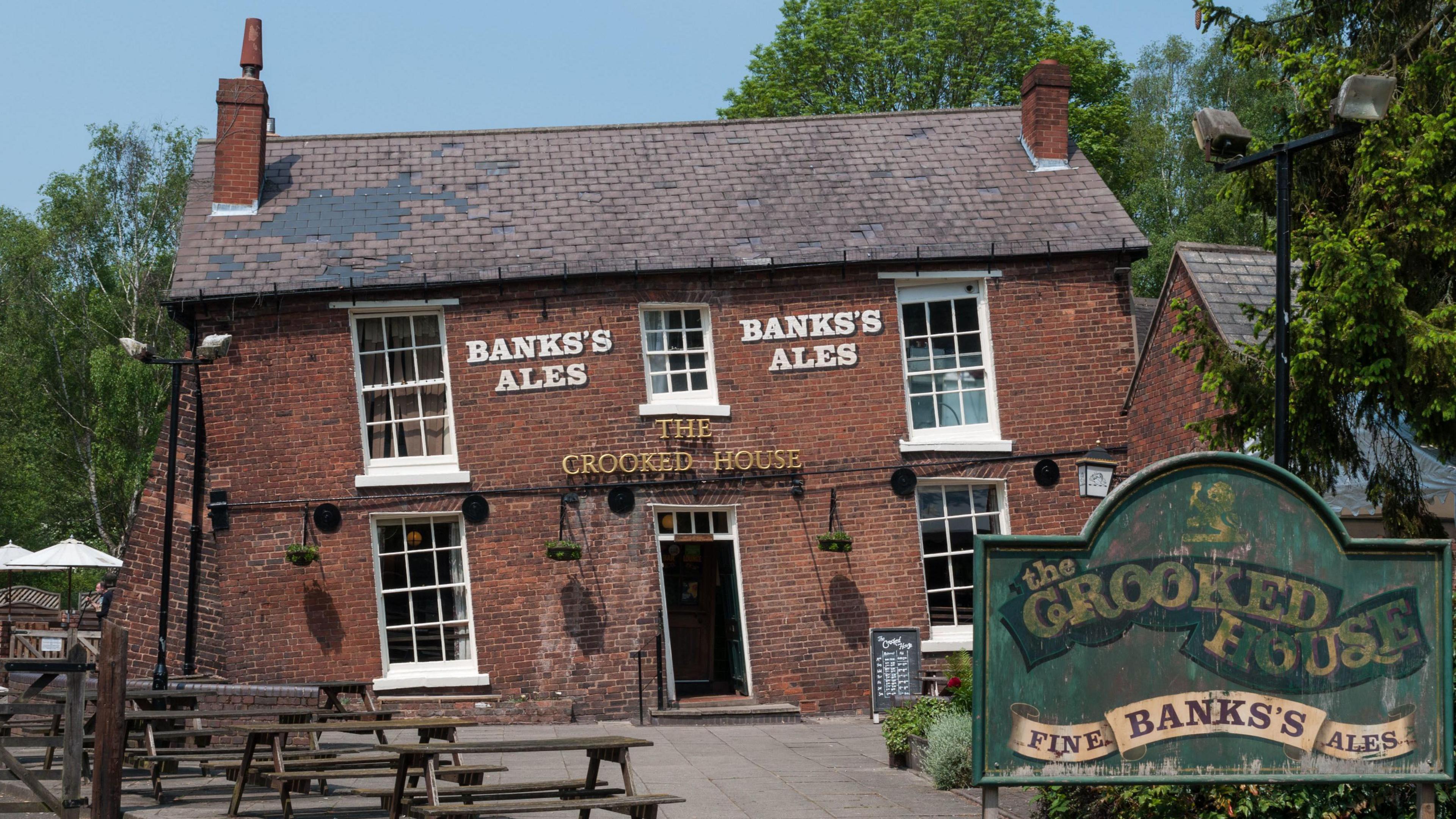 The front of The Crooked House pub, which is crooked due to subsidence causing one side of the pub to partially sink. The Crooked House sign is over the front door in gold letters with Banks's Ale twice at the top of the building in white letters. A green sign in front of the pub says "The Crooked House - Fine Banks's Ales".