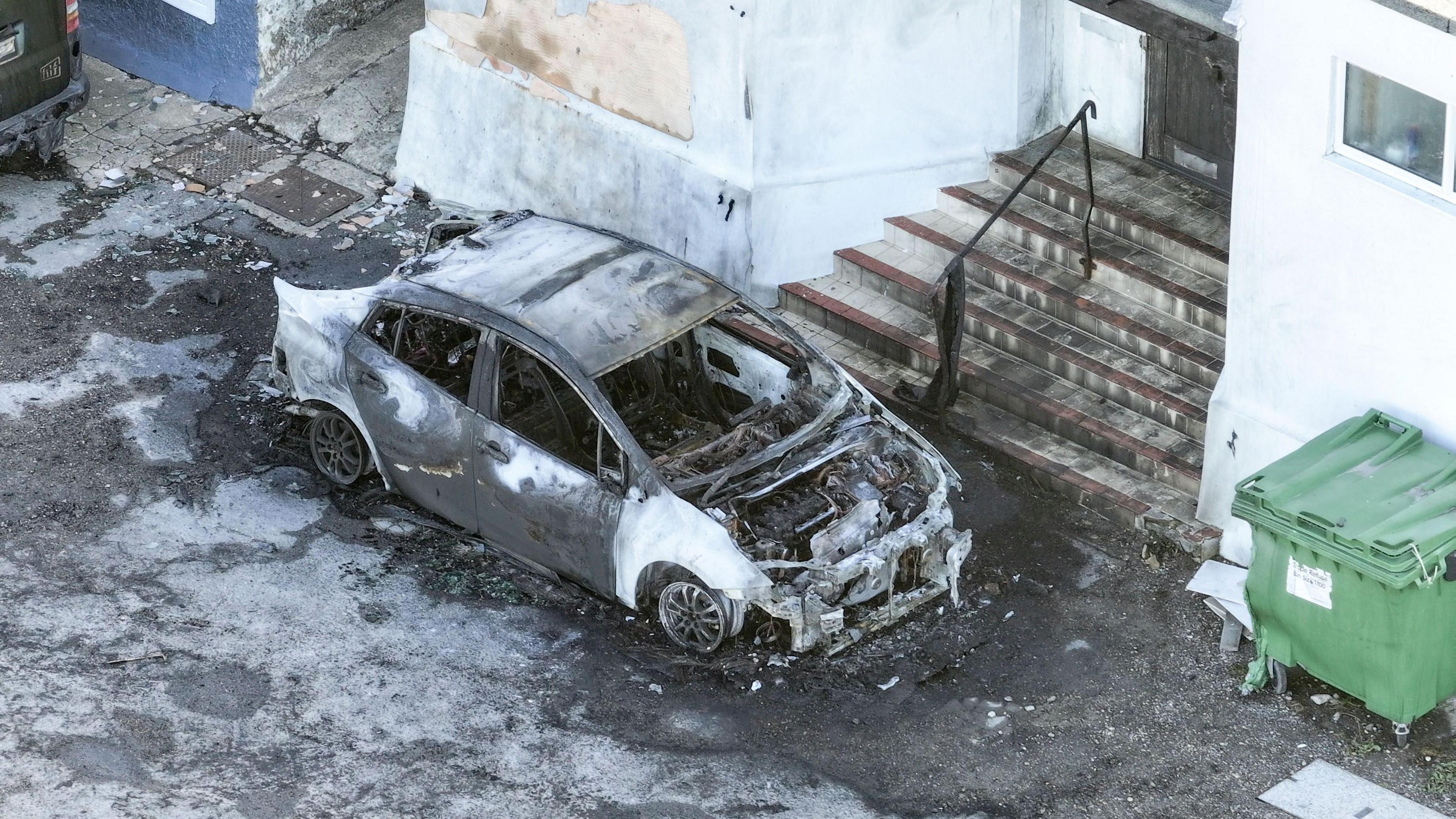 A scorched car outside the mosque. It is parked in front of the entrance steps, which are covered in black soot and have partially melted railings.
