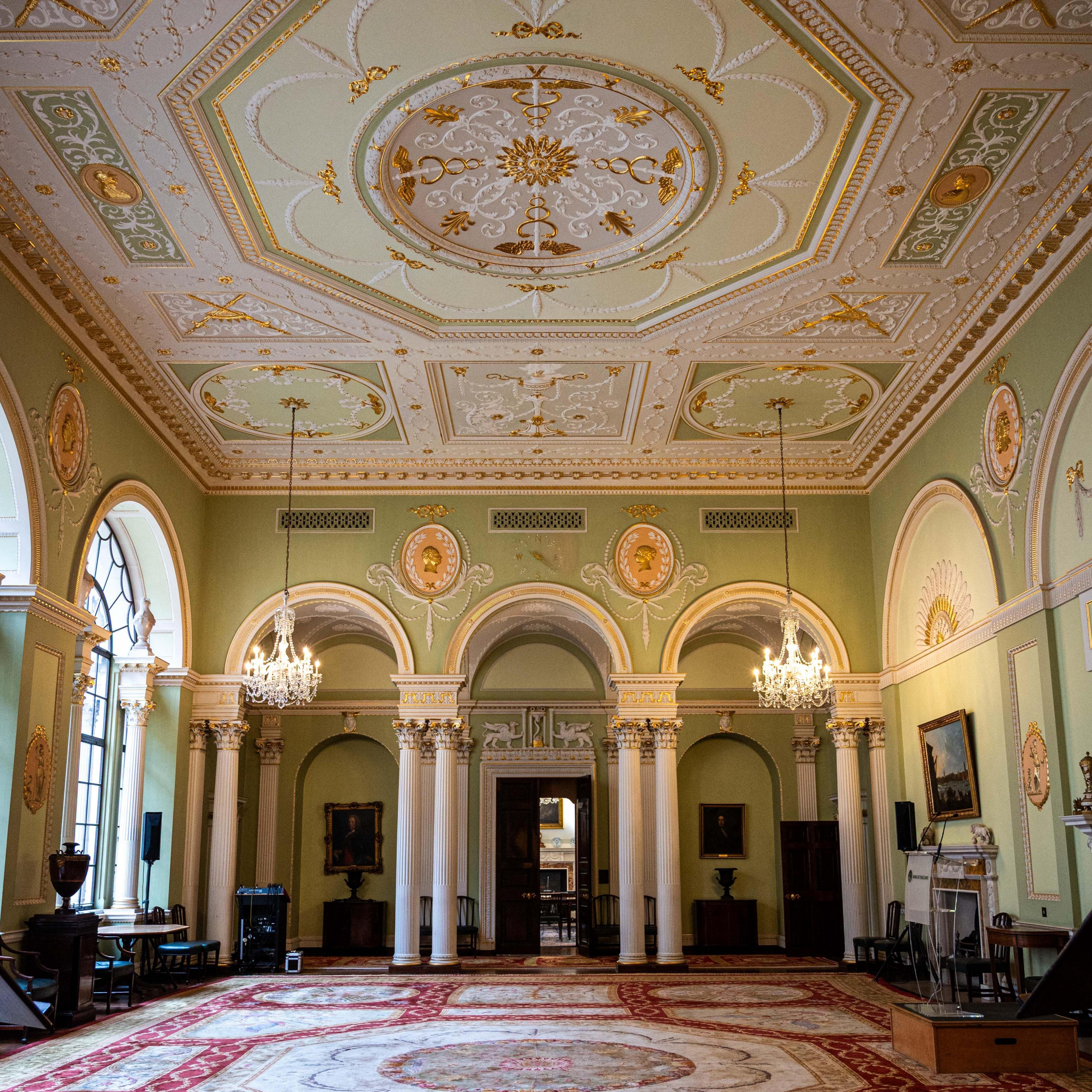 The court room in the Bank of England which has decorated green walls, an elaborate gold, green, white and gold ceiling and a patterned rug with white columns and arches on the far side of the room