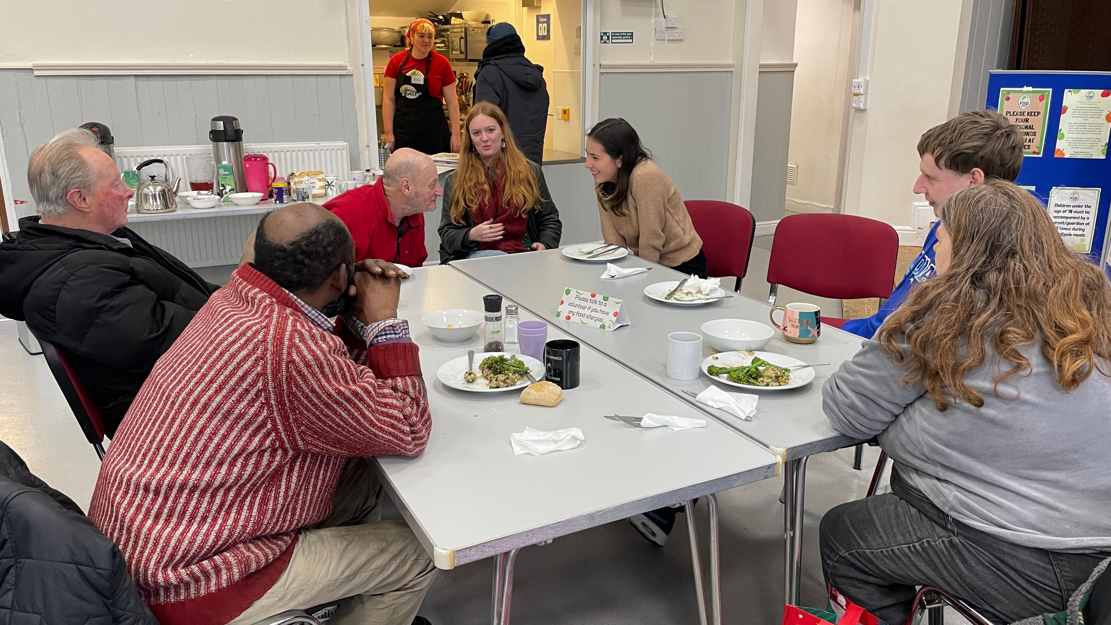 A group of people sit around a dining table and talk. There is crockery and plates of food scattered across the table


