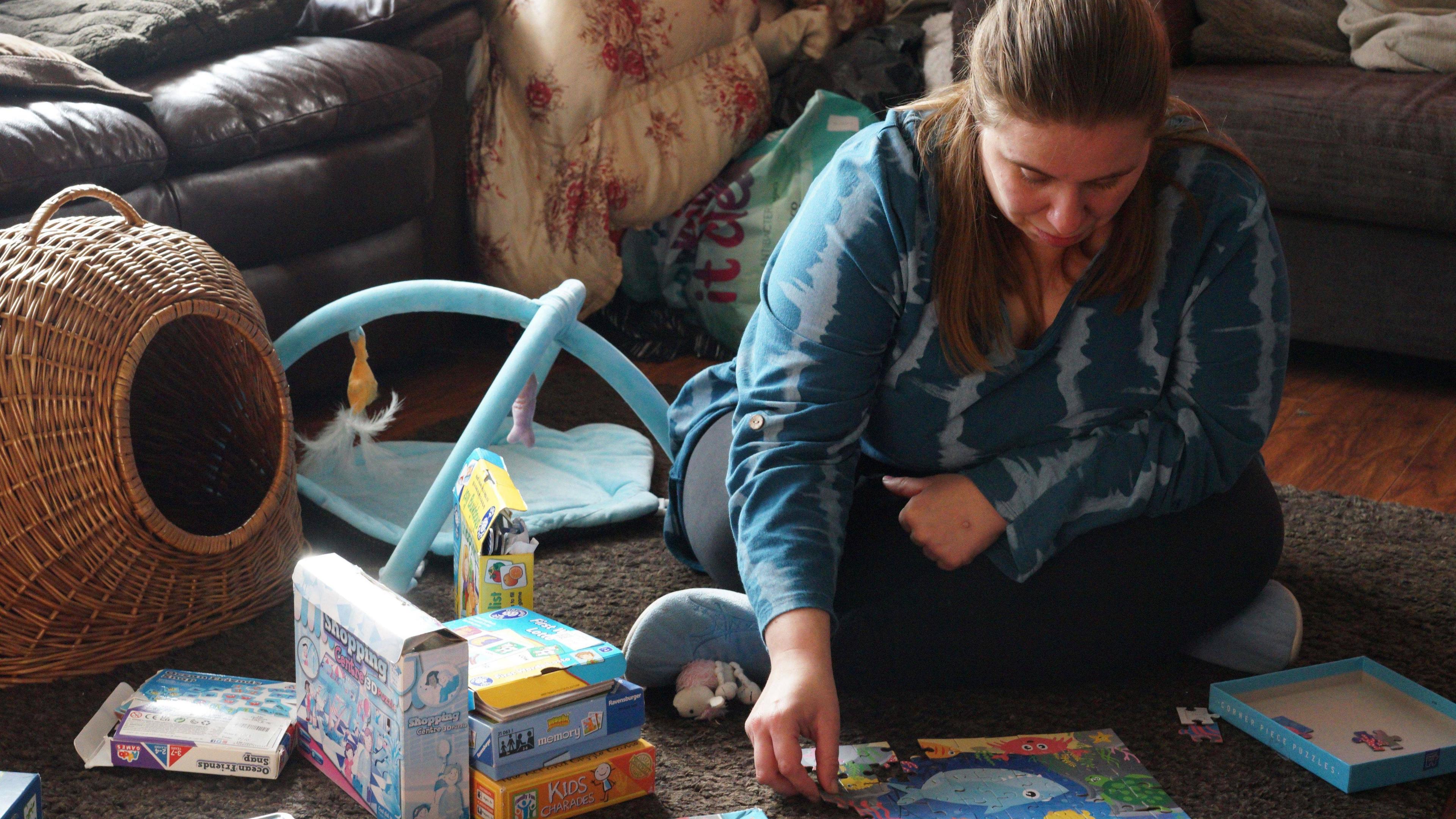 Dawn is sitting on the living room floor doing a puzzle, she is surrounded by the sofas, some cat toys and boxes of puzzles.