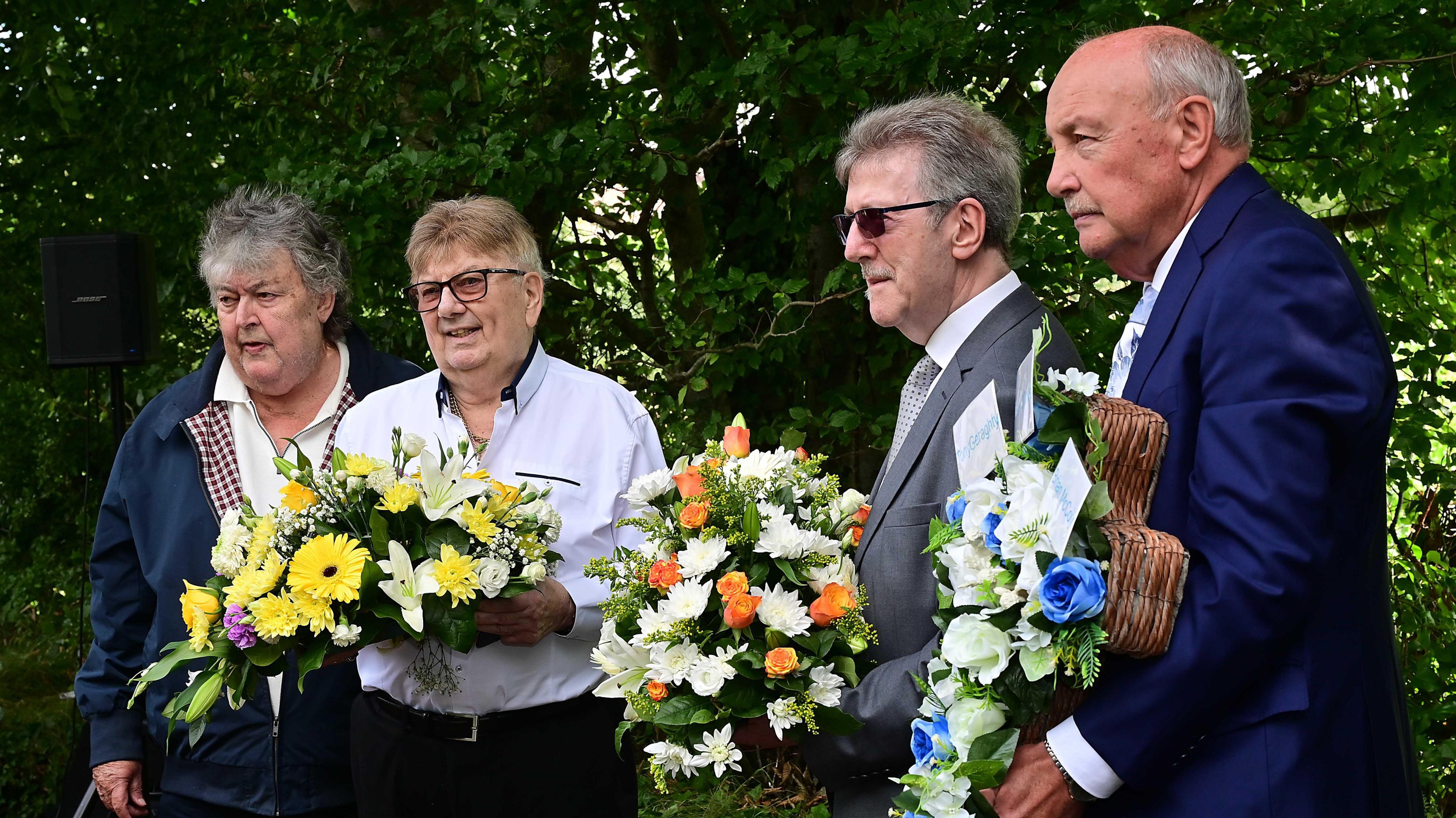 Four men standing together. Three of the men are holding flowers.