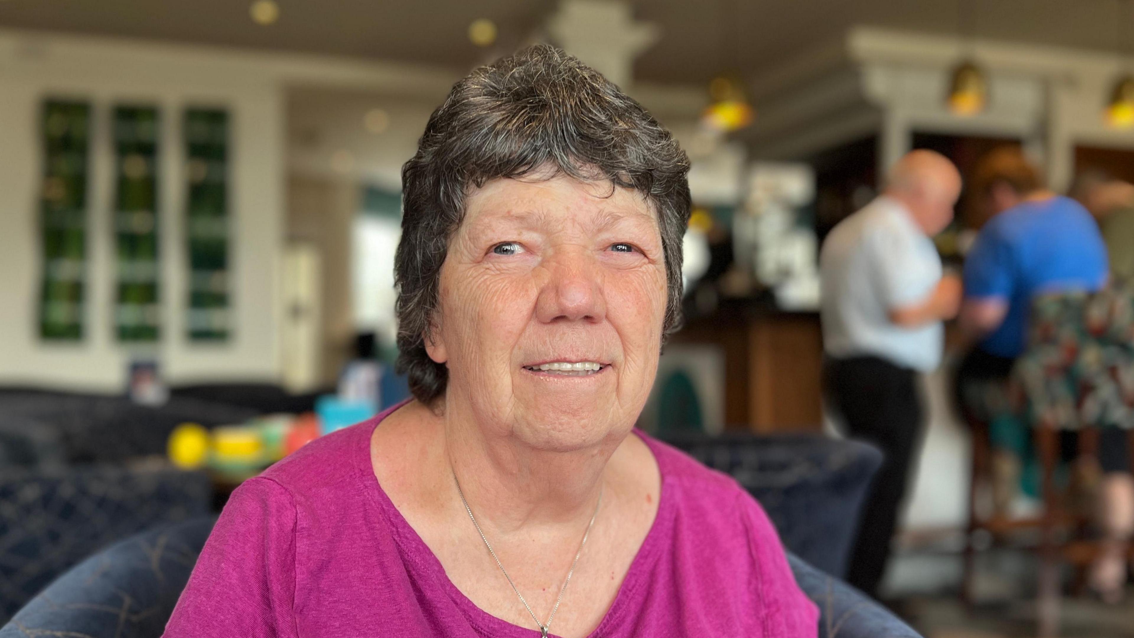 Marilyn Bland sat in a chair facing the camera in the bar room of the hotel She is wearing a pink top with a silver chain. The background is blurred, but several people can be seen standing at the bar.