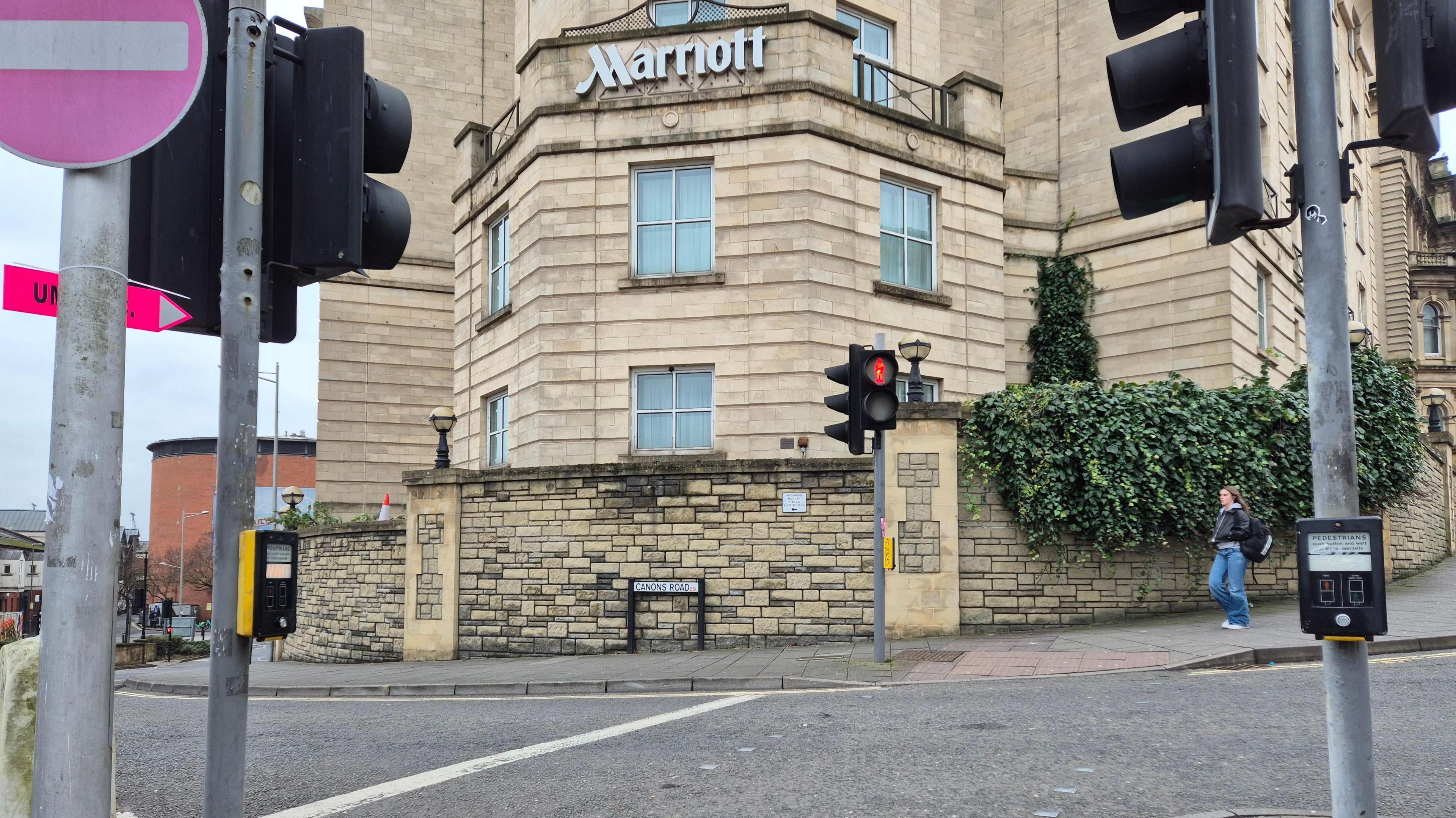 The pedestrian crossing on Anchor Road outside the Marriott Hotel in Bristol. The crossing is empty, but a pedestrian approaches from up the hill. The hotel sign is clear to see behind the retaining way. Several sets of traffic lights are also in shot. 