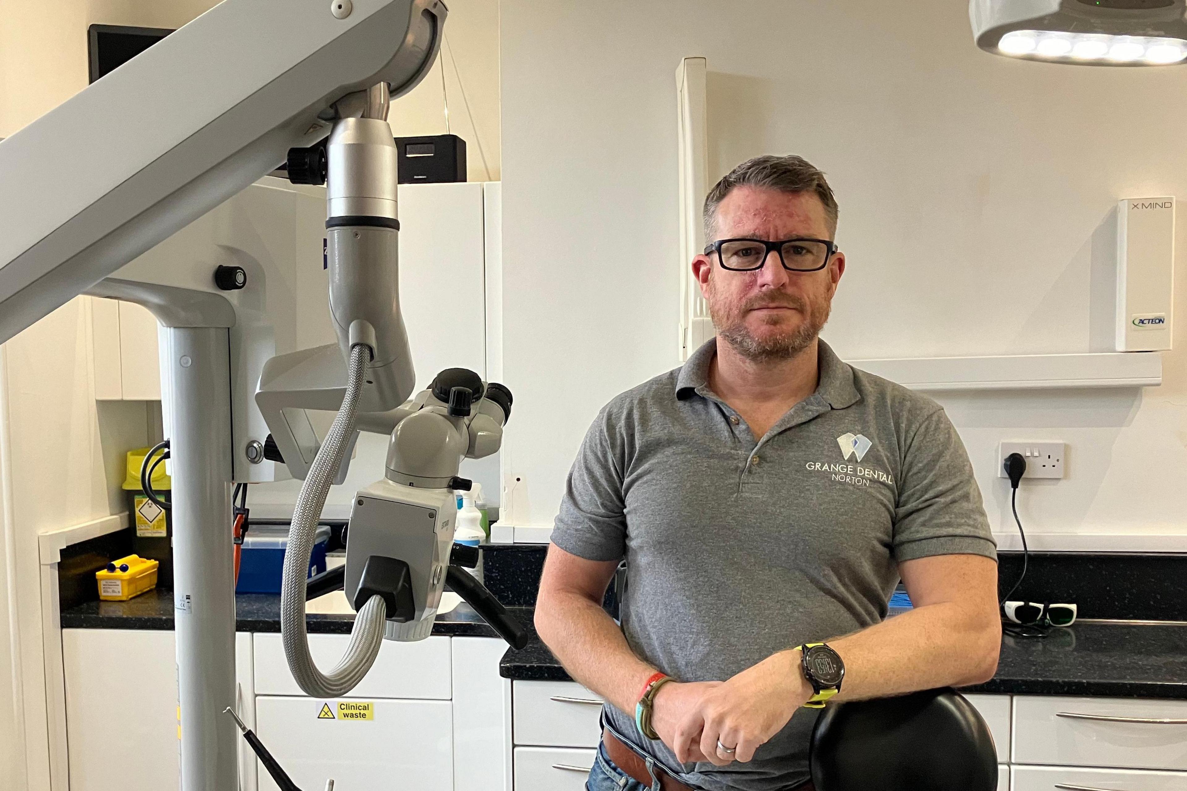 Paul Woodhouse of the British Dental Association is standing in a dentist's operating room, leaning on the back of the chair in which patients would usually sit. He has brown hair and glasses and is wearing a t-shirt with a logo for Grange Dental Practice. Dentist's equipment is to his right.