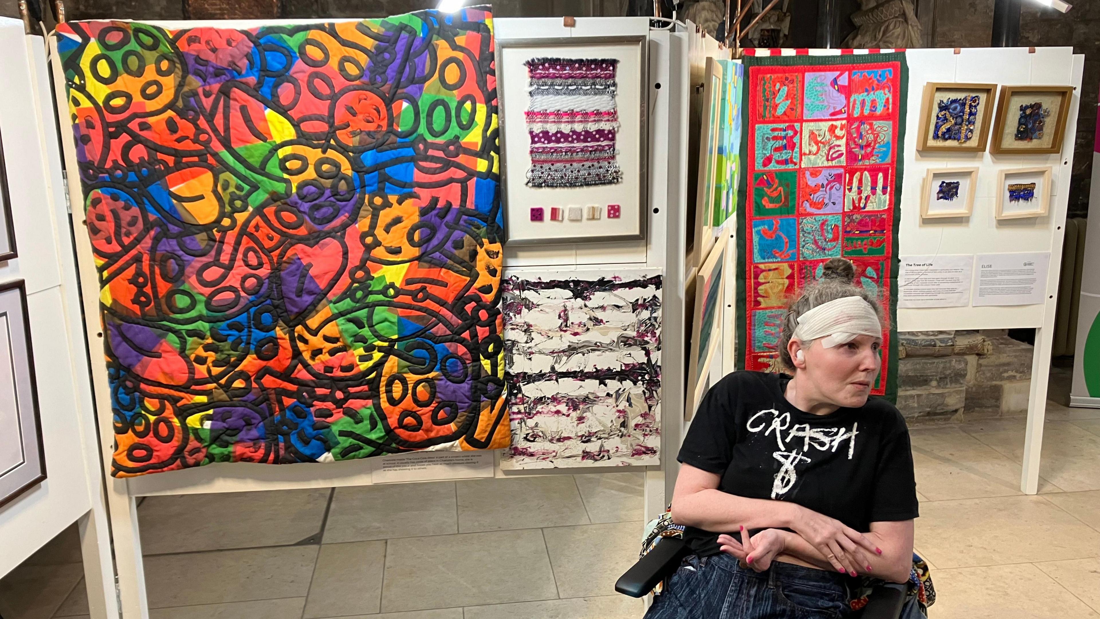 An artist in a wheelchair sat in front of a colourful piece of artwork at an exhibition at St John the Baptist Church in Cirencester