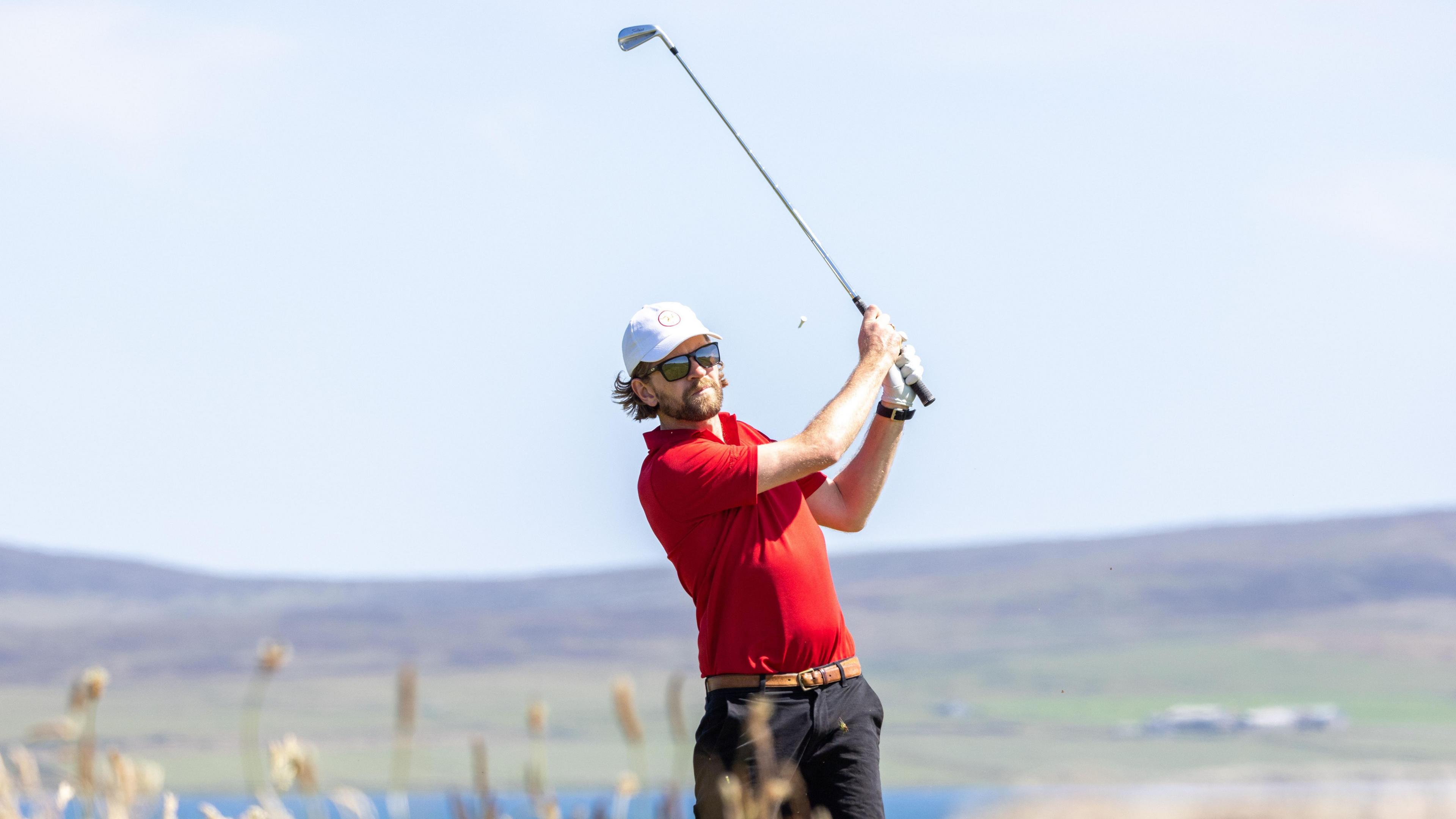 Daryl Callister wears a red T-shirt and dark trousers, with a white cap and dark sunglasses. He swings a silver golf club high over his shoulder with water and low hills in the background.