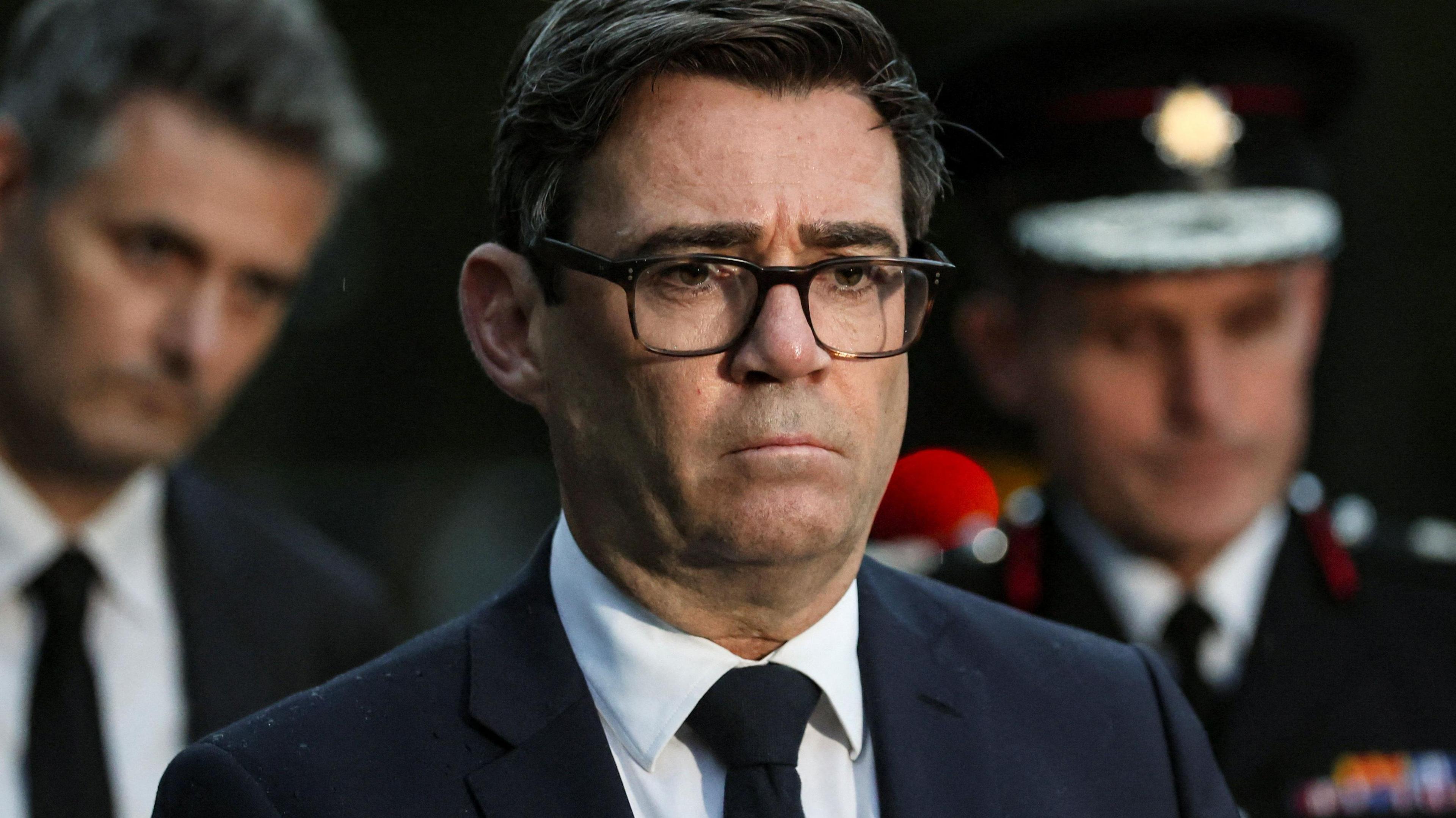 Mayor of Greater Manchester Andy Burnham with brown hair wearing black glasses and a black suit. He looks sombre.