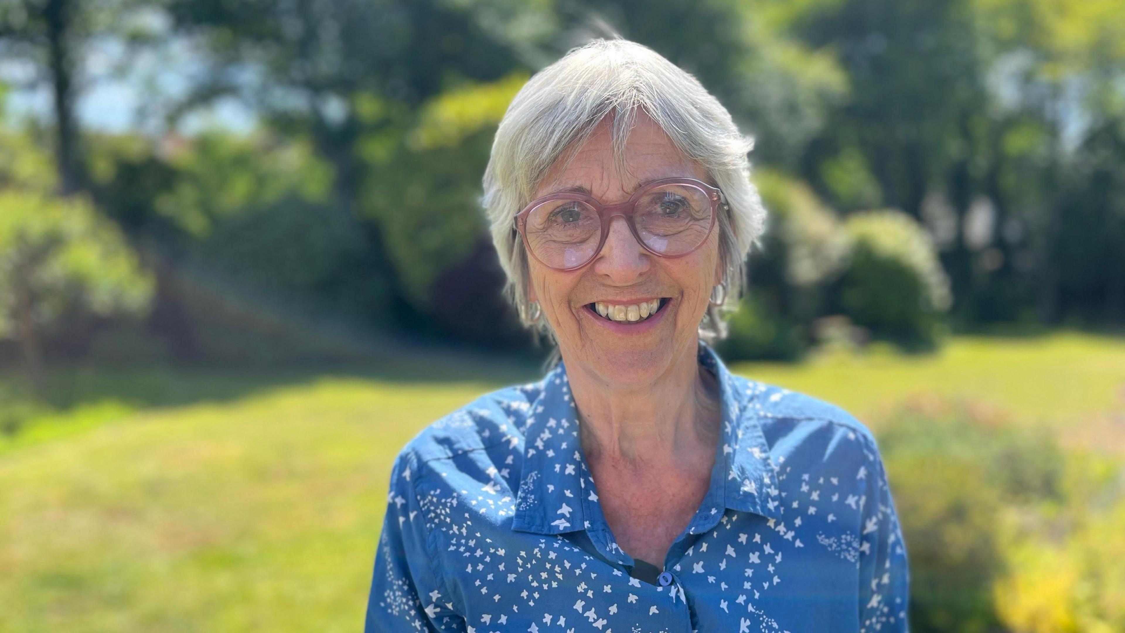 An older woman with short hair, a blue shirt and glasses stands in a field smiling on a sunny day.