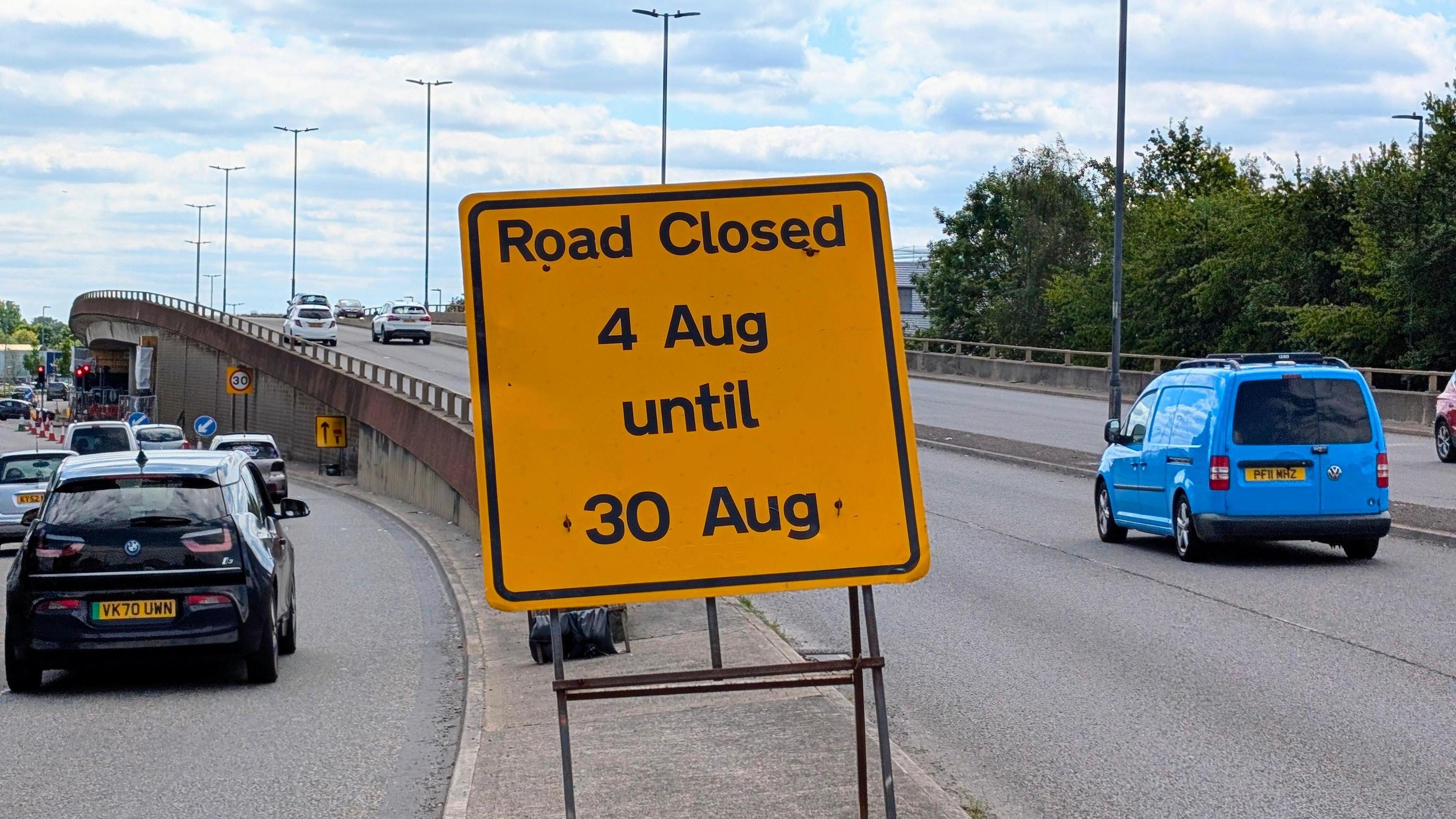 A yellow sign saying road closed 4 Aug until 30 Aug. There is a flyover in the distance with cars driving down the road.