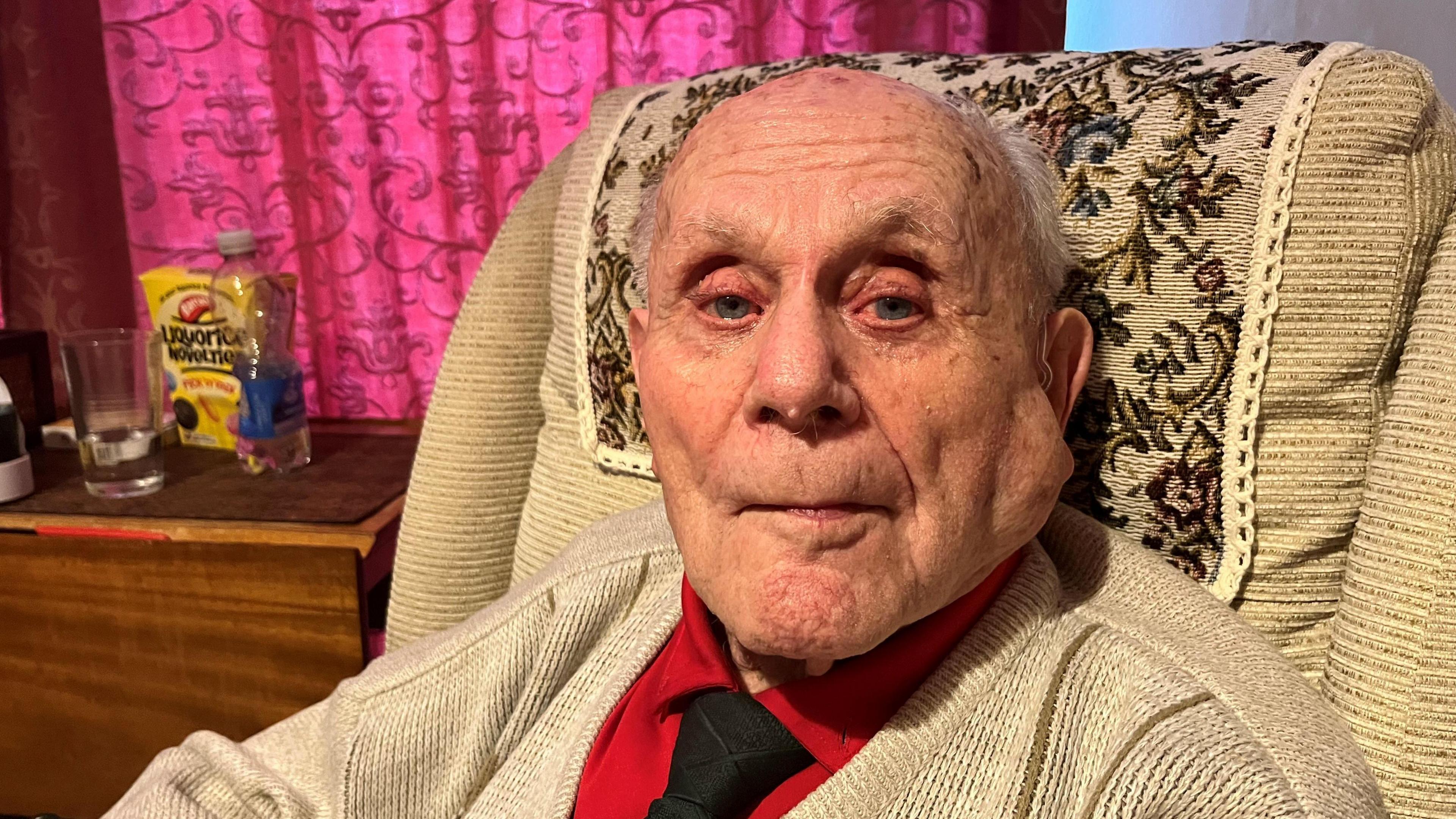Stanley Eastgate, 102, has very thin grey hair on the sides of his head he has a red shirt with black tie under a beige cardigan and is sitting in a wing chair with a small table beside him with a glass and bottle of water on and some liquorice novelties in a yellow box.