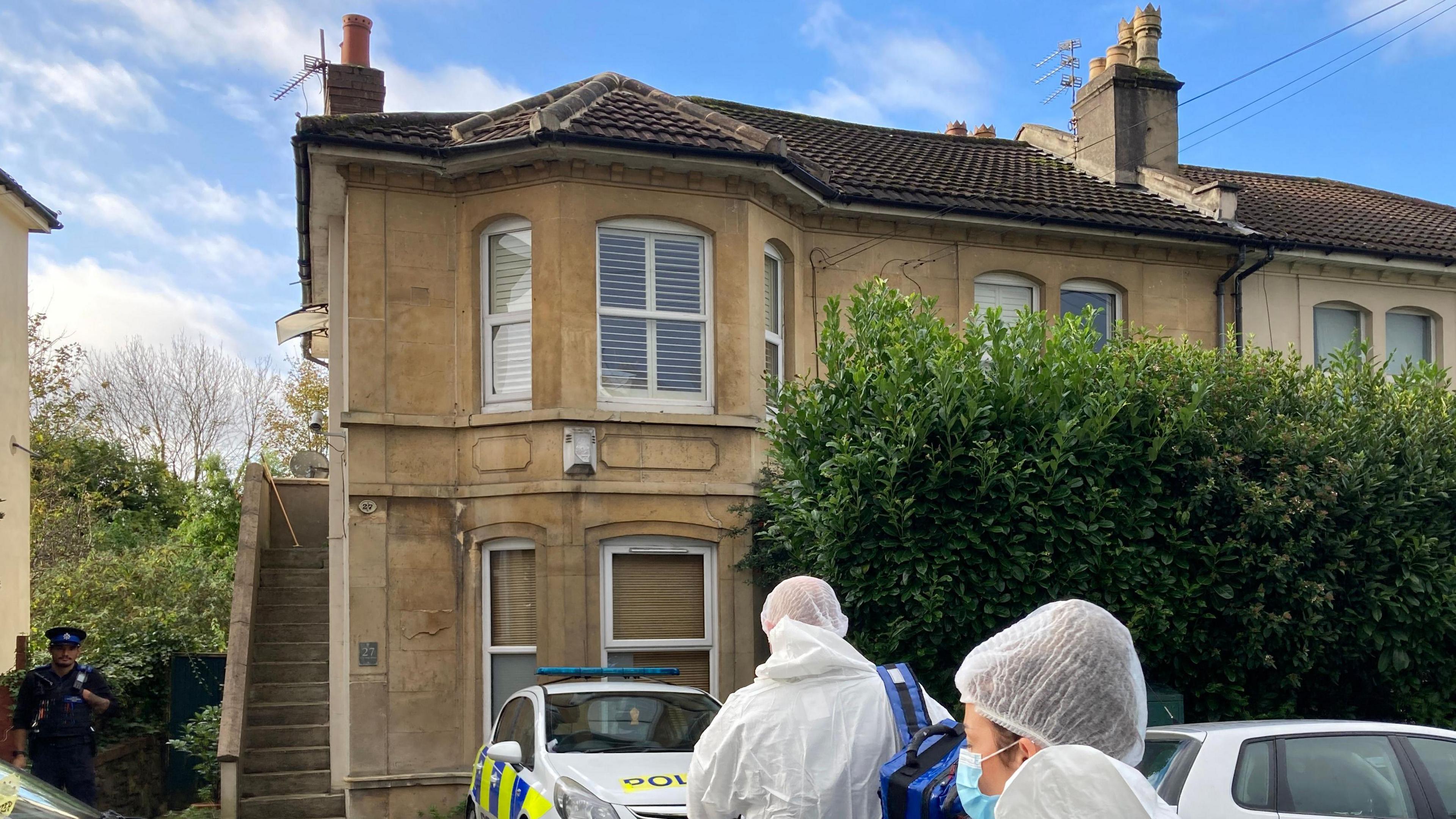 Two forensic workers in white suits walking to cream-coloured house with police cars outside