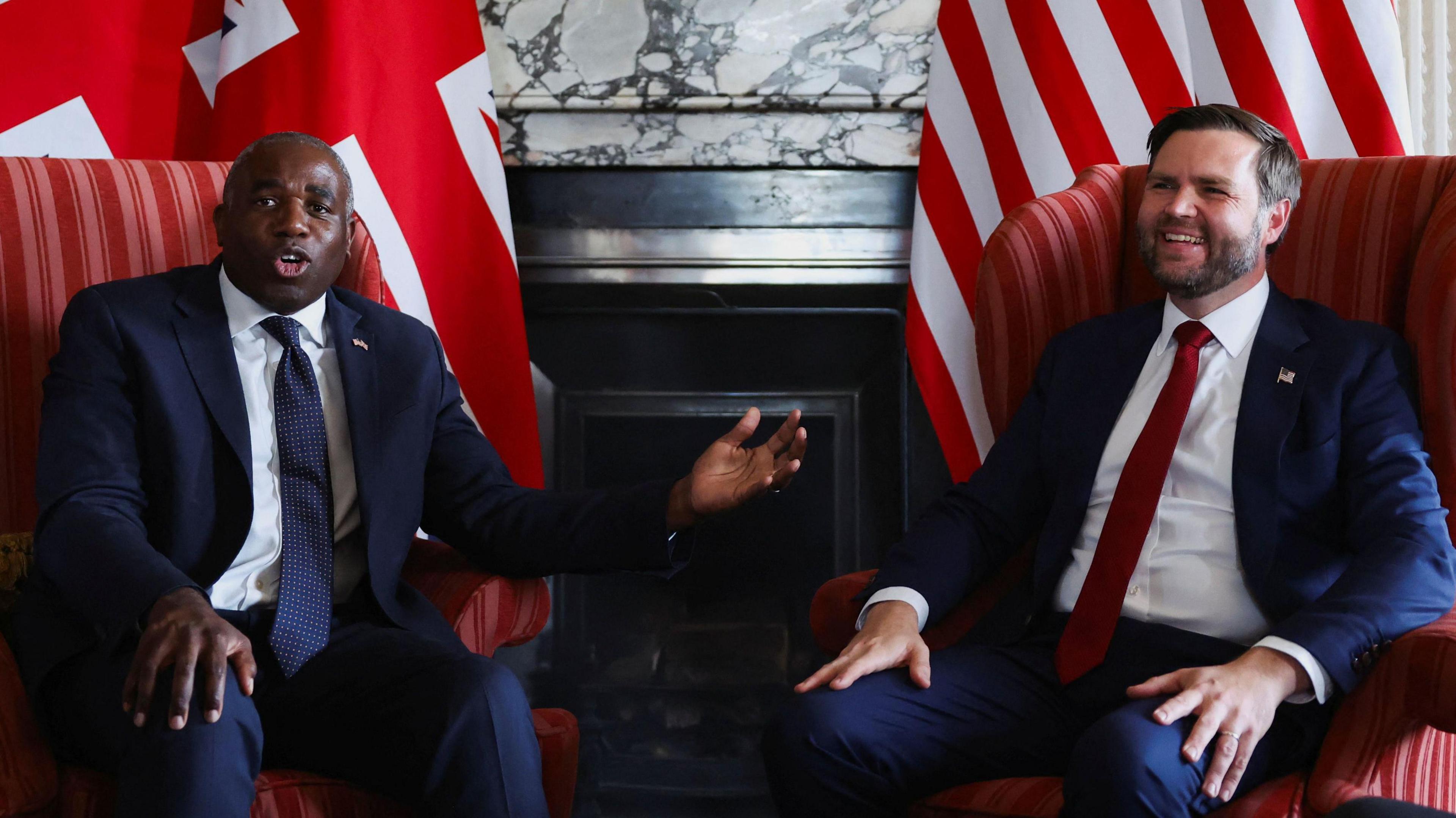 David Lammy, wearing a navy suit and tie, gestures to JD Vance, who is wearing a navy suit and red tie, and has a relaxed smile, as they sit in armchairs during a photo call in front of a British flag and an American flag at Chevening
