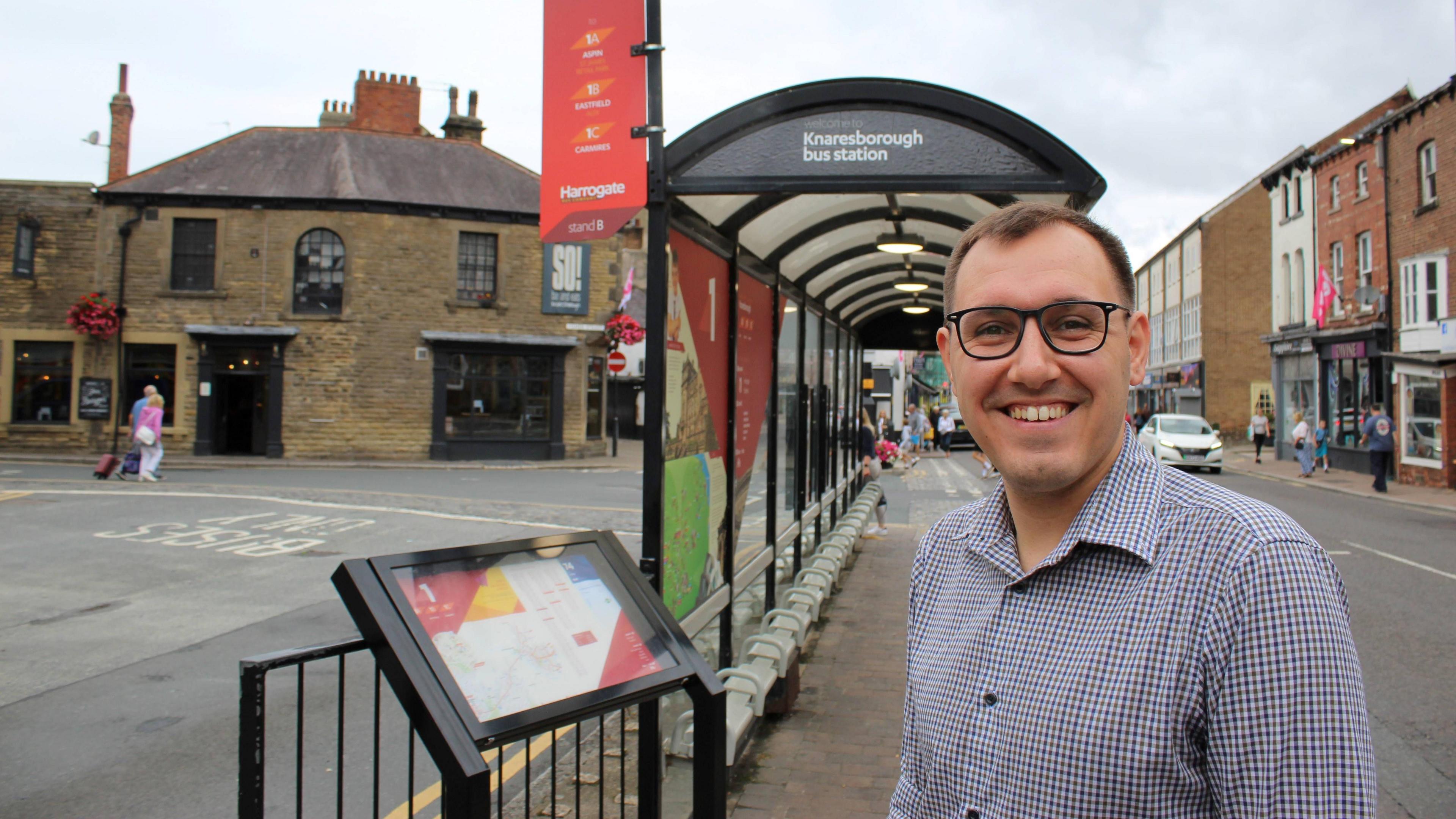 A man with short brown hair is wearing glasses and a blue and white checked shirt and smiling. He is stood at a bus interchange.