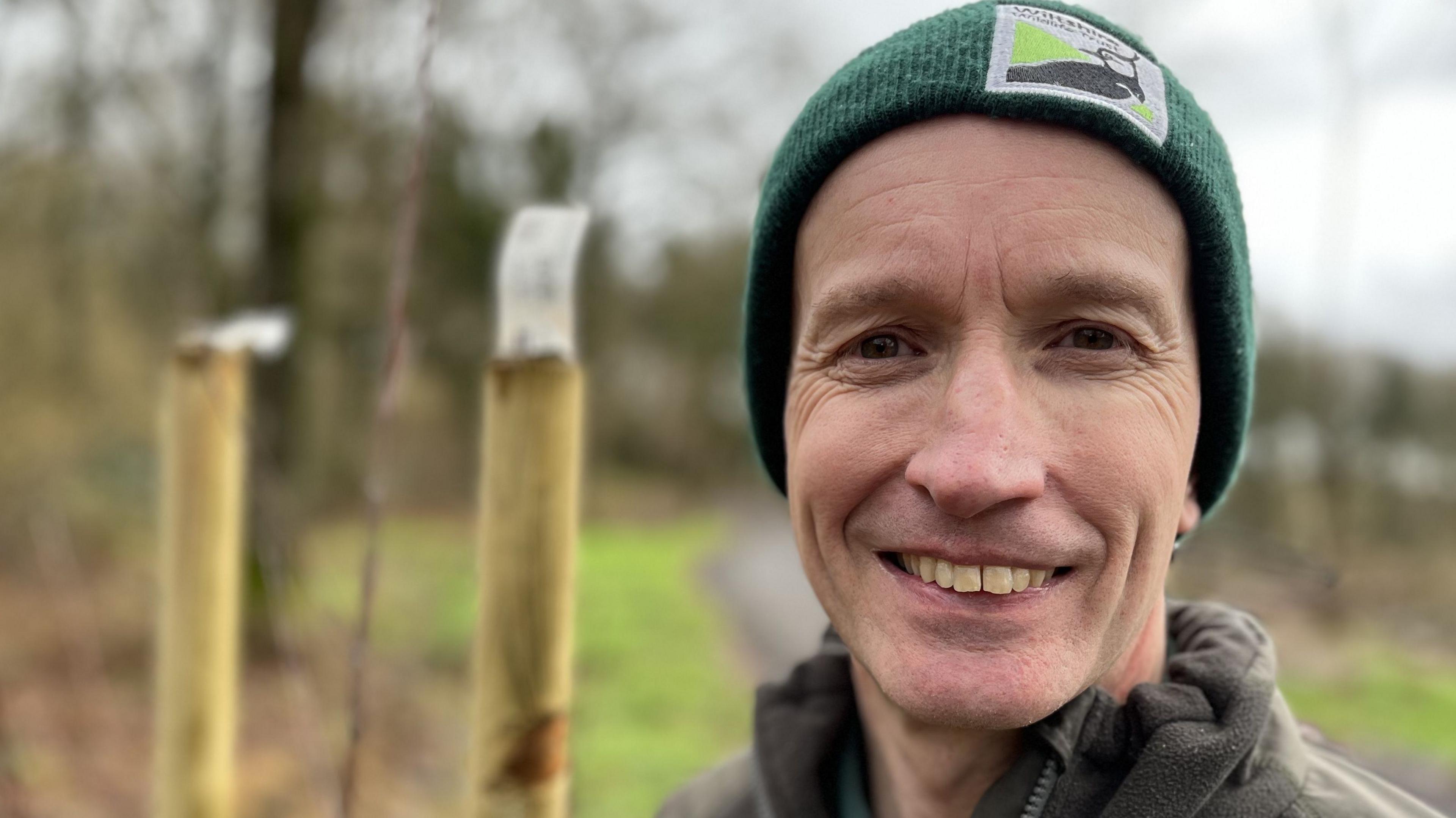 James Ravine in a Wiltshire Wildlife Trust beanie hat stood next to saplings inside tree guards