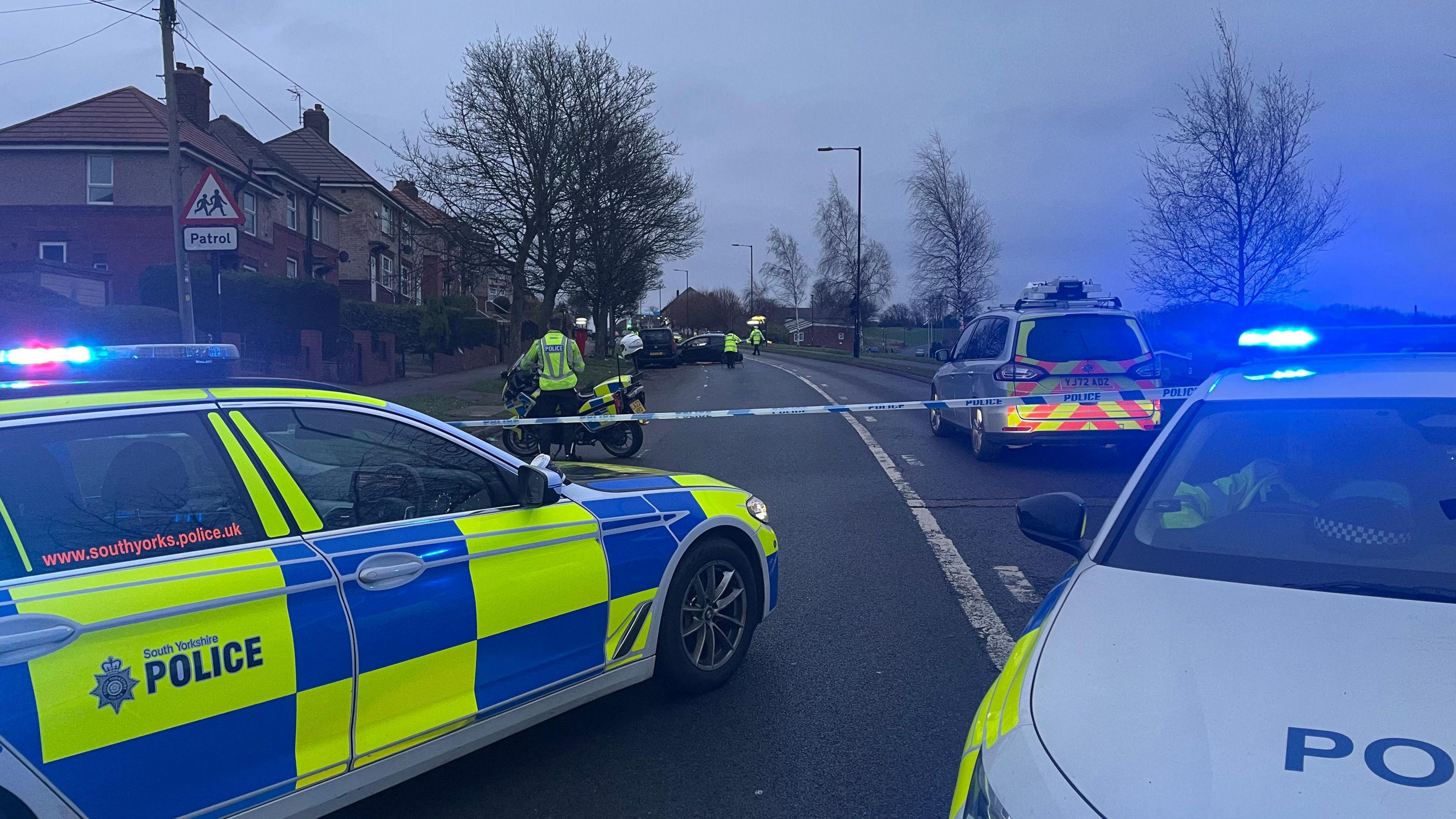 Three police cars, a police motorbike and several officers at the scene of a road crash. Blue flashing lights are in shot, with the highway taped off to allow for investigation work. A crashed car can be seen in the distance. 