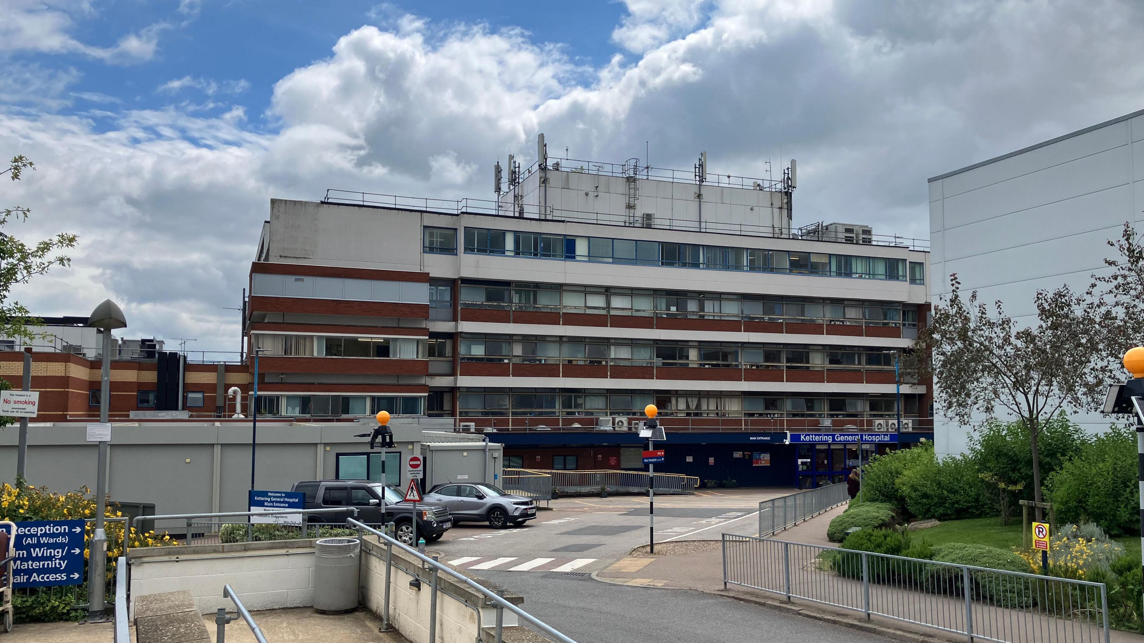 A photo of Kettering General Hospital, a multi-storey building which looks run down. There is construction site in the foreground and a number of cars parked besides a zebra crossing. 