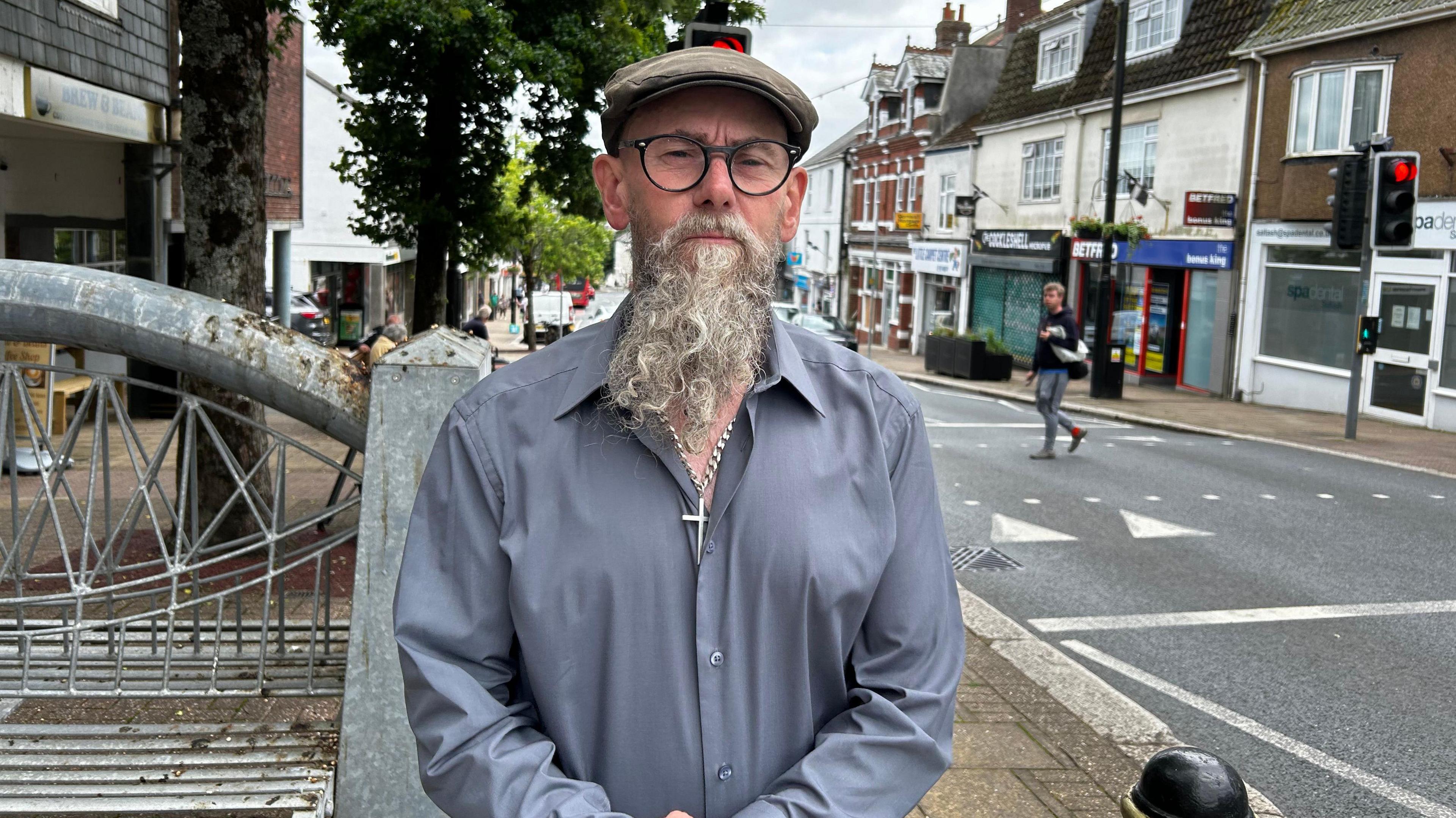 A man with a long grey beard, wearing a blue shirt and a brown cap and glasses. He is looking directly at the camera. Behind him to the right is a road and traffic lights. To the left of him is the high street and a metal bench.