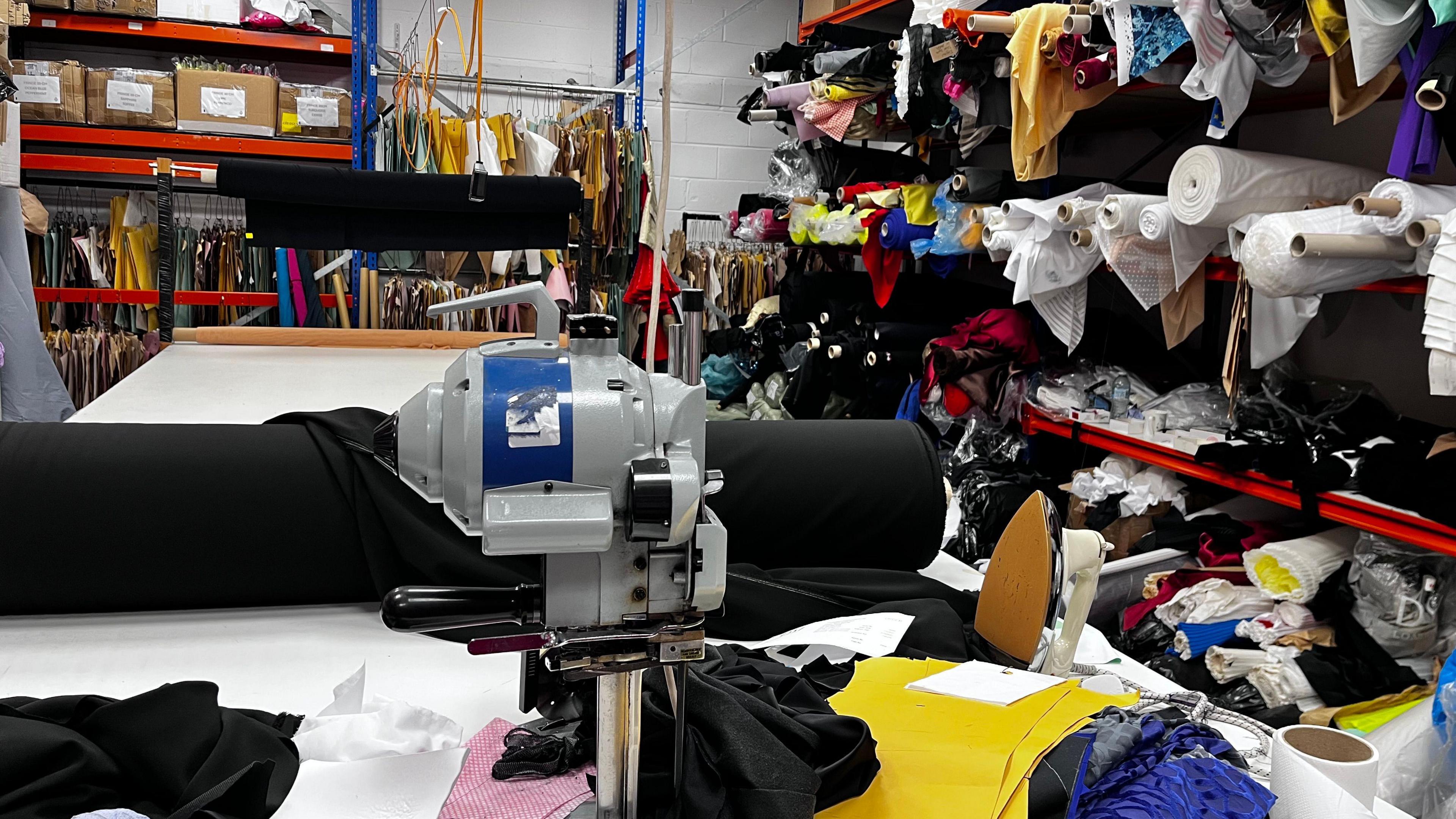 A sewing machine on a table, with piles of fabric in the background on shelves in a warehouse.