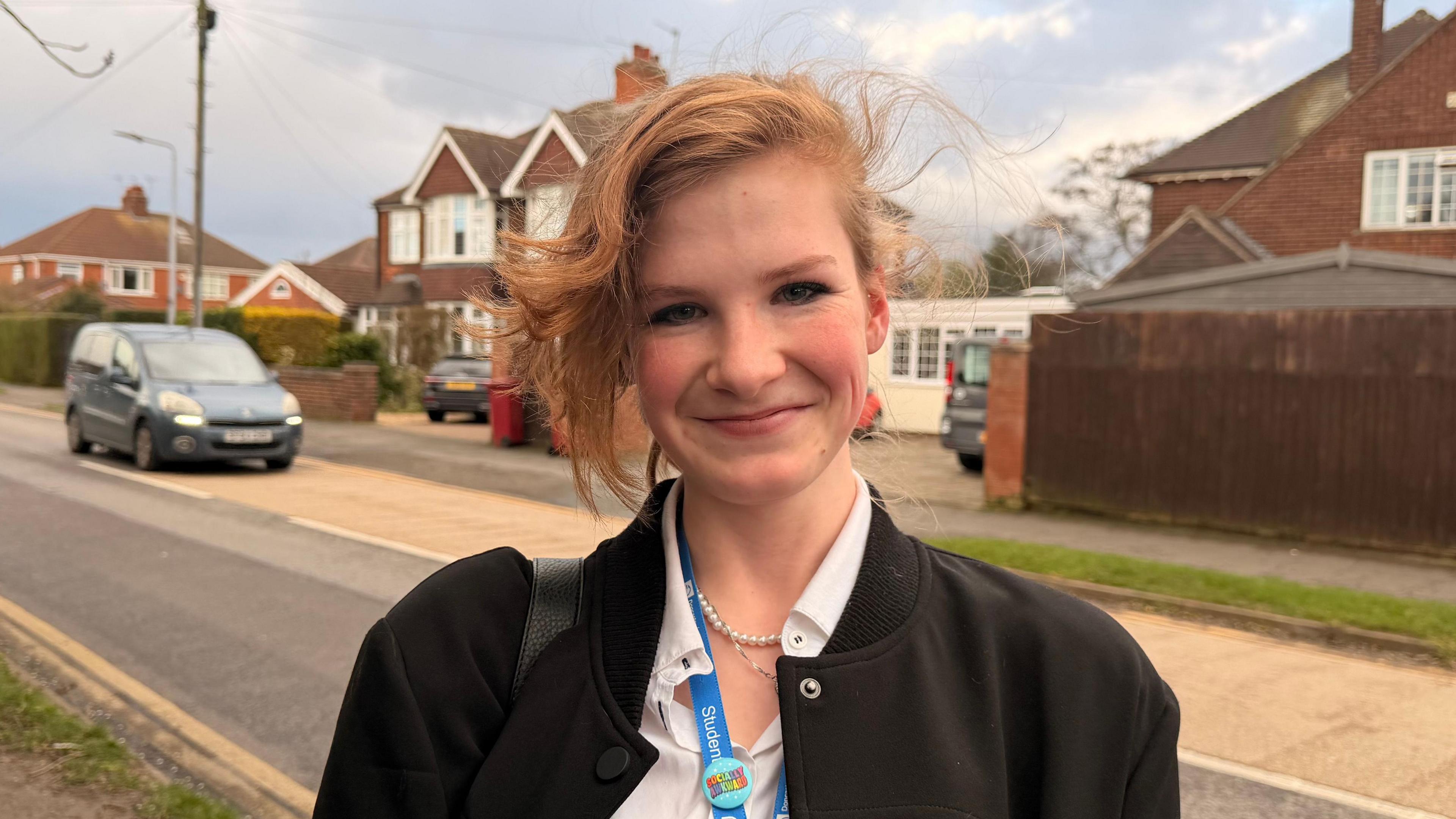 A young woman smiles with closed lips as she stands in a residential street with red-brick semi-detached houses behind her. She has ginger hair in an up-do with side parting. She is wearing a black coat with a white shirt, pearl necklace and blue lanyard. 