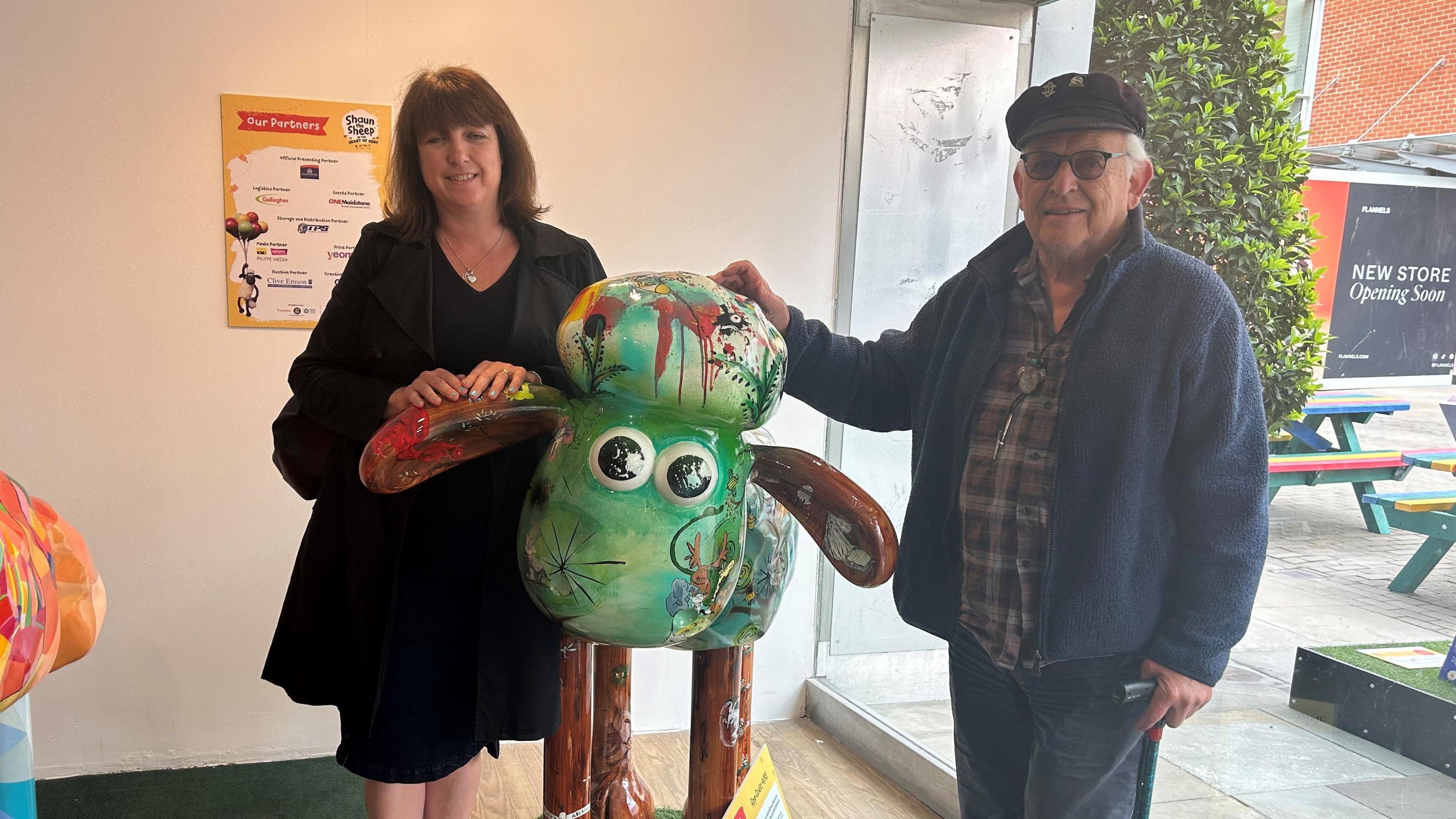 Two people stood next to a green painted cartoon Shaun the Sheep sculpture. On the left a woman wearing a black dress and the right a man wearing a checked shirt with a cap. 