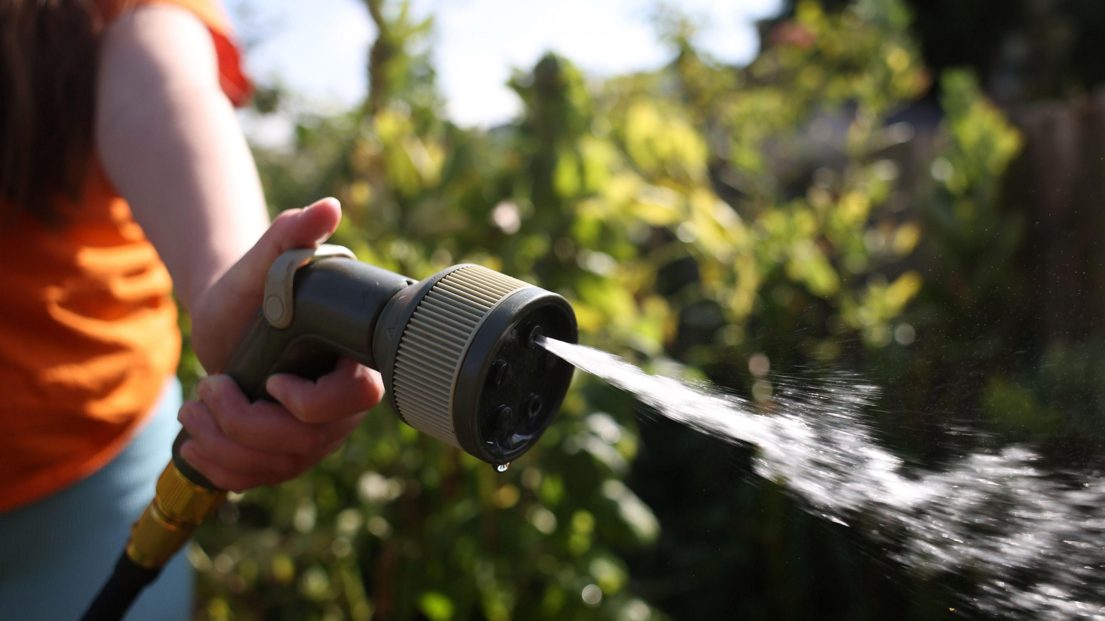 A close up image of a sprinkler hose, with 5 water holes, and water spraying out of one of them. A person's hand is clasped around the hose, and you can see their arm and orange T-shirt.