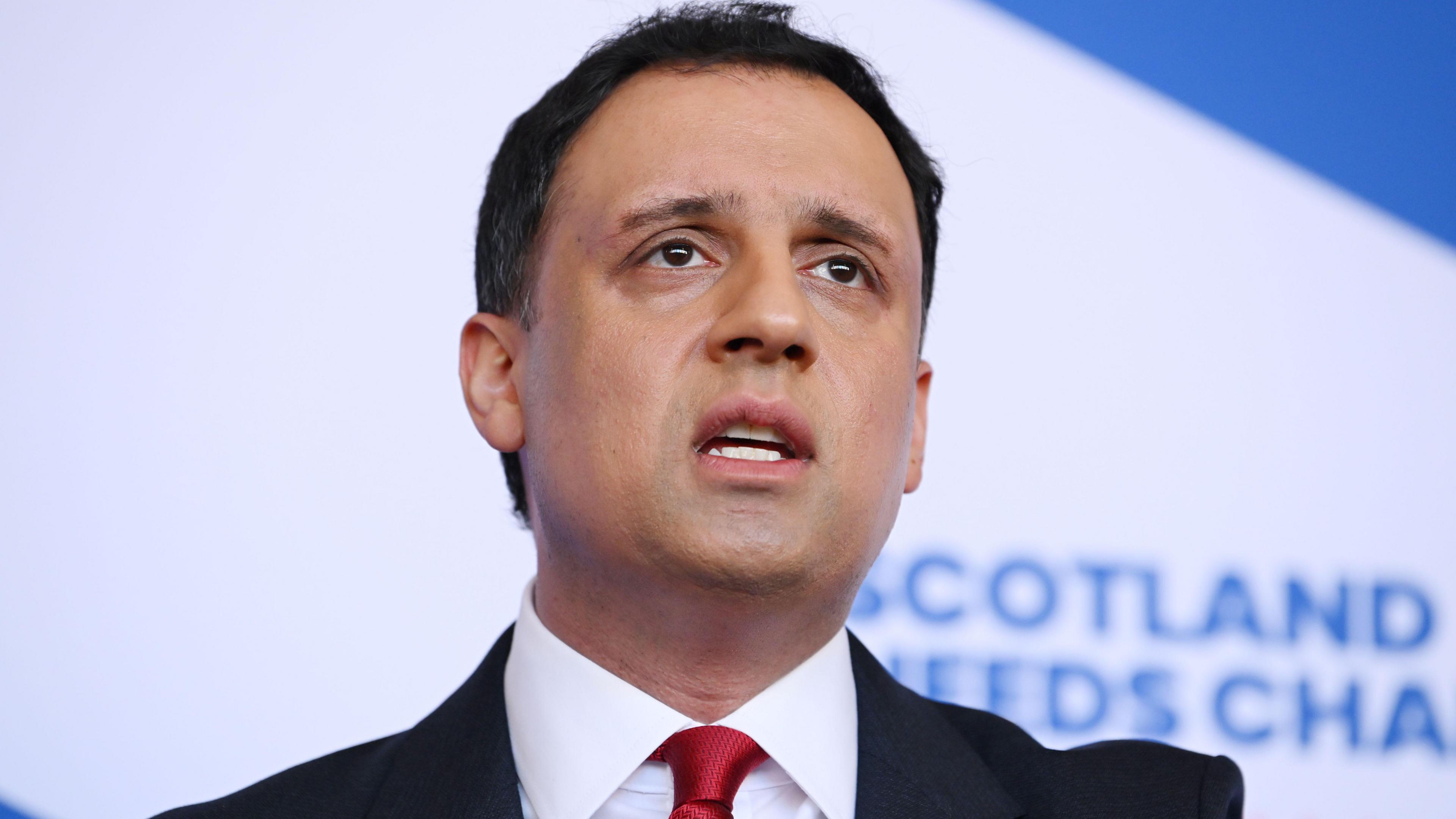 Anas Sarwar po shikon me ashpërsi kamerën. Ai ka flokë të shkurtër e të errët dhe mban veshur një xhaketë kafe dhe një këmishë të bardhë. Pas tij ka aktivistë dhe shtëpi.