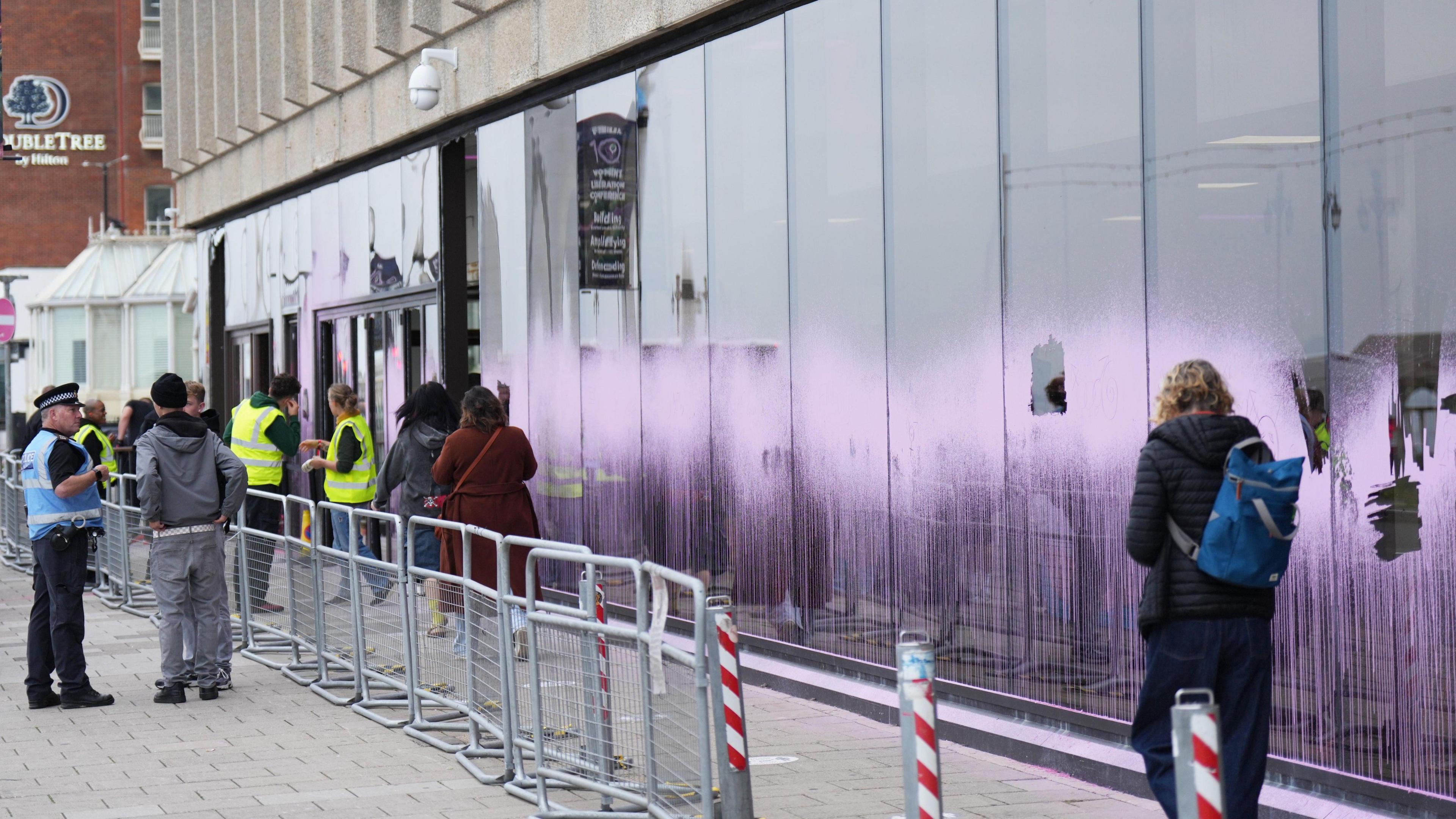 A long window at the front of a building is spray-painted pink. Some panes have been smashed.