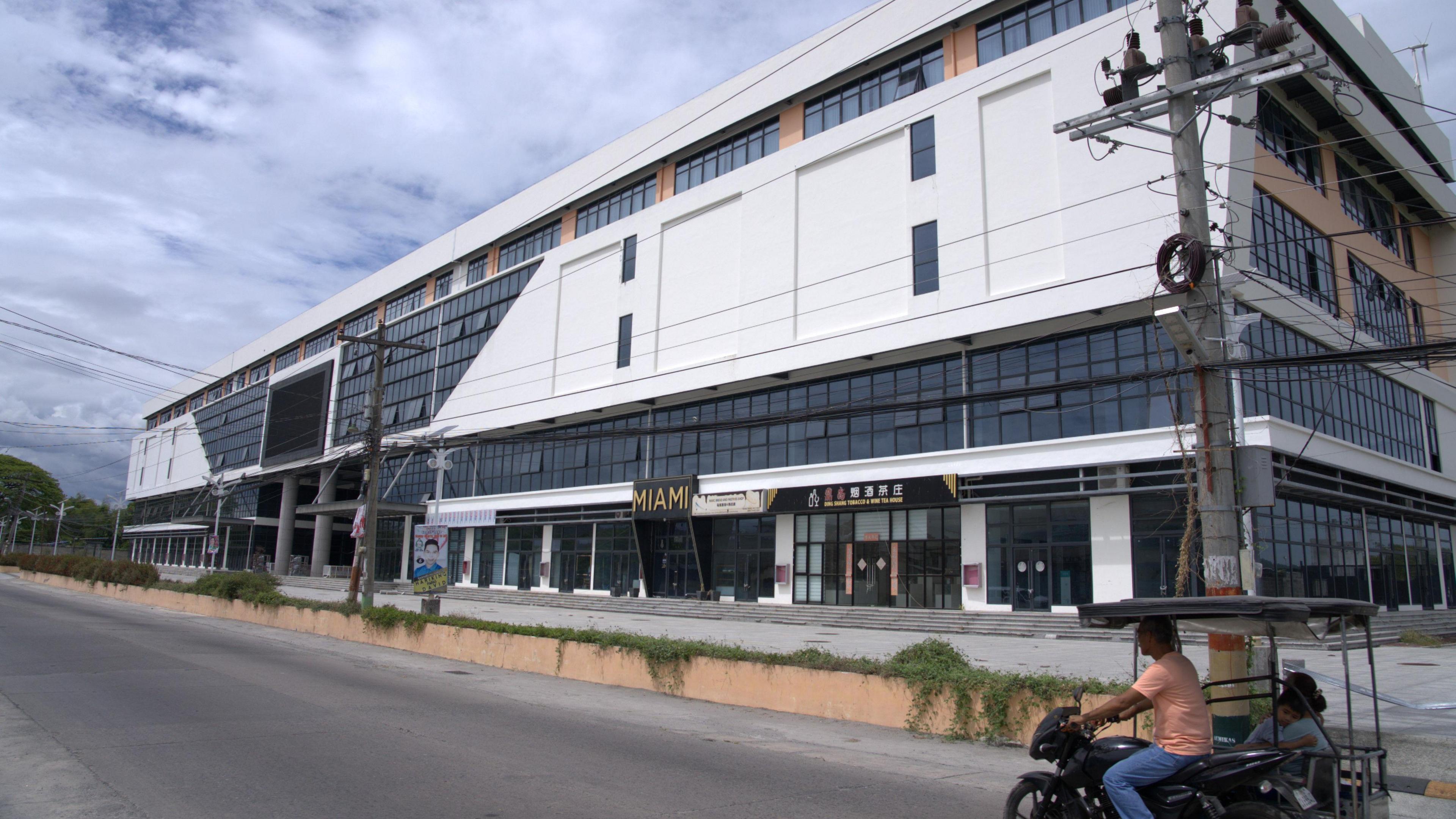 A long, low building faced in white panels and dark blue coloured glass. It has multiple floors, and features an arcade of businesses on ground level. An archway has been driven through the centre of the building, resting on two large pillars.