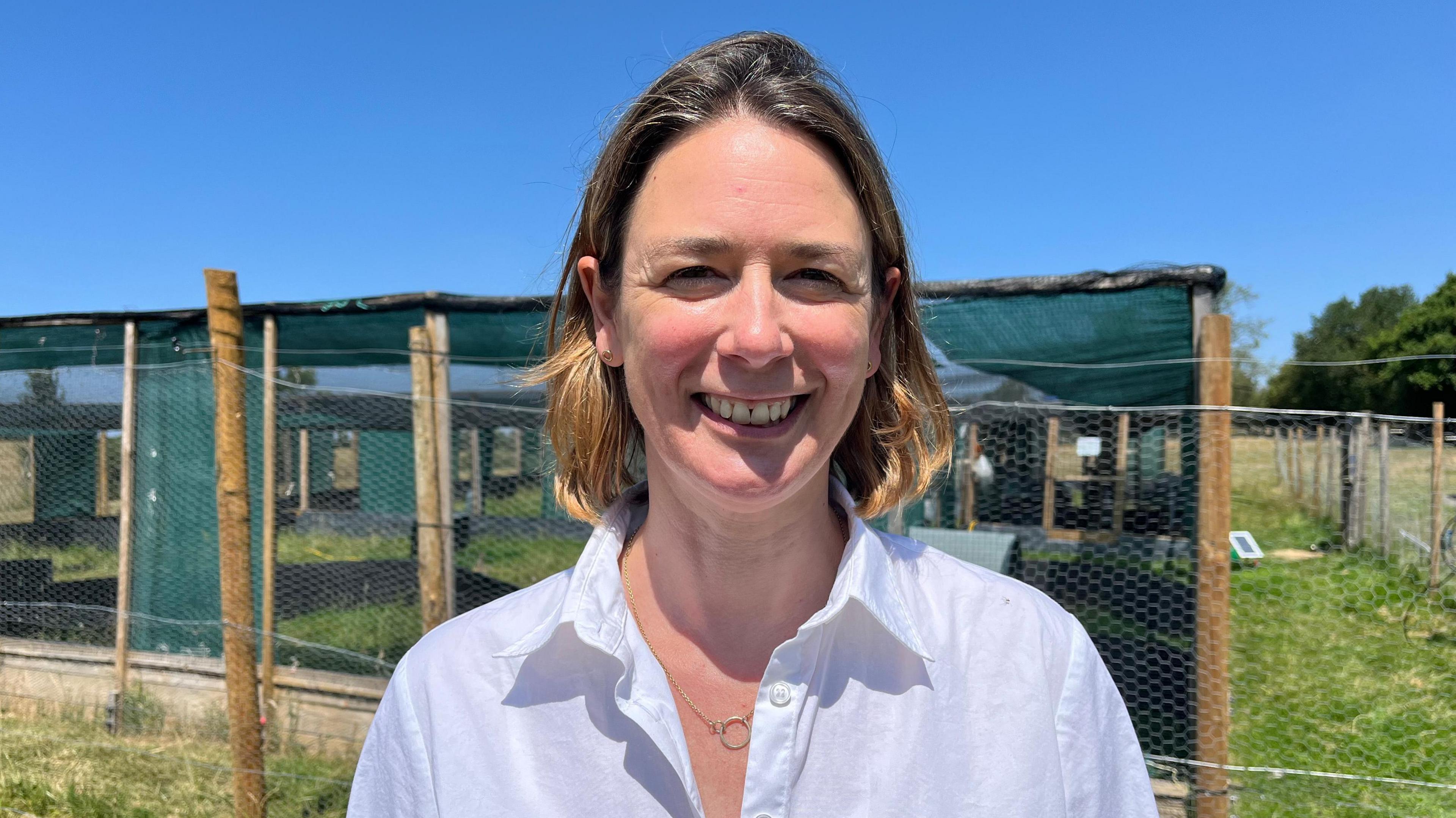 Parker photographed looking into the camera and smiling, wearing a white collared shirt. She is stood on a large grassy field, with pens for the curlews behind her built out of chicken wire, wooden posts, and green material. The sky is clear and blue and there is strong sunshine. 