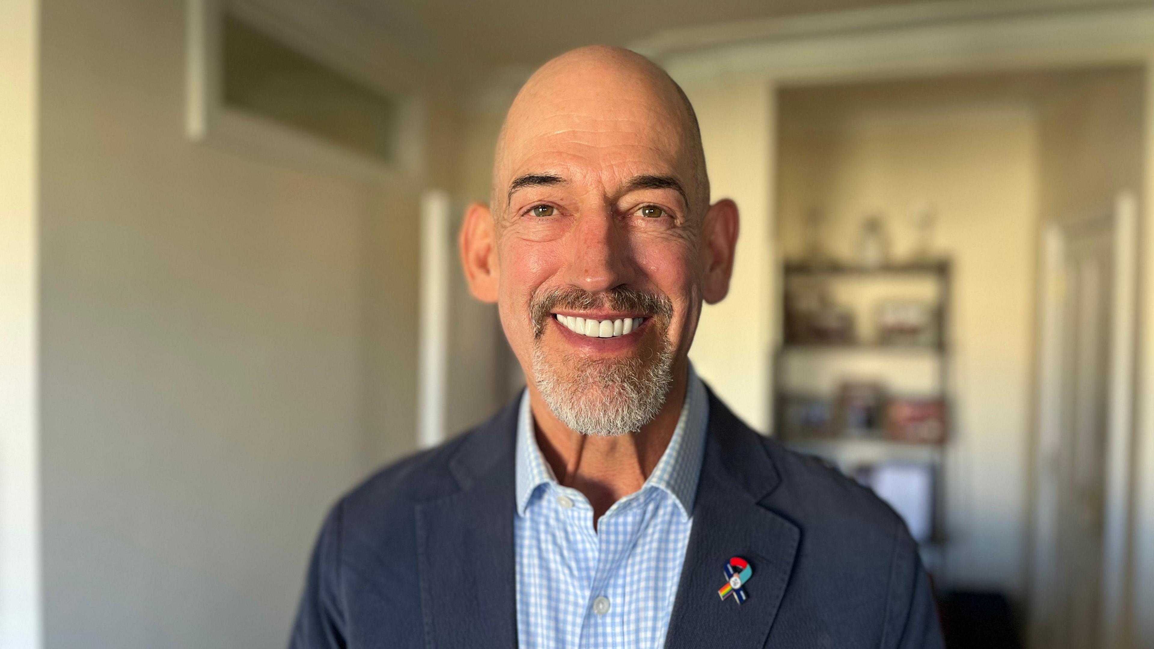 Roly Woods wearing a pale blue and white checked shirt and navy blazer, he has a special badge from the Navy which has a Pride flag on it. He is bald and has a beard. He is smiling at the camera. The background is blurred, but appears to be his home, with a shelving unit covered in items visible but blurred behind him