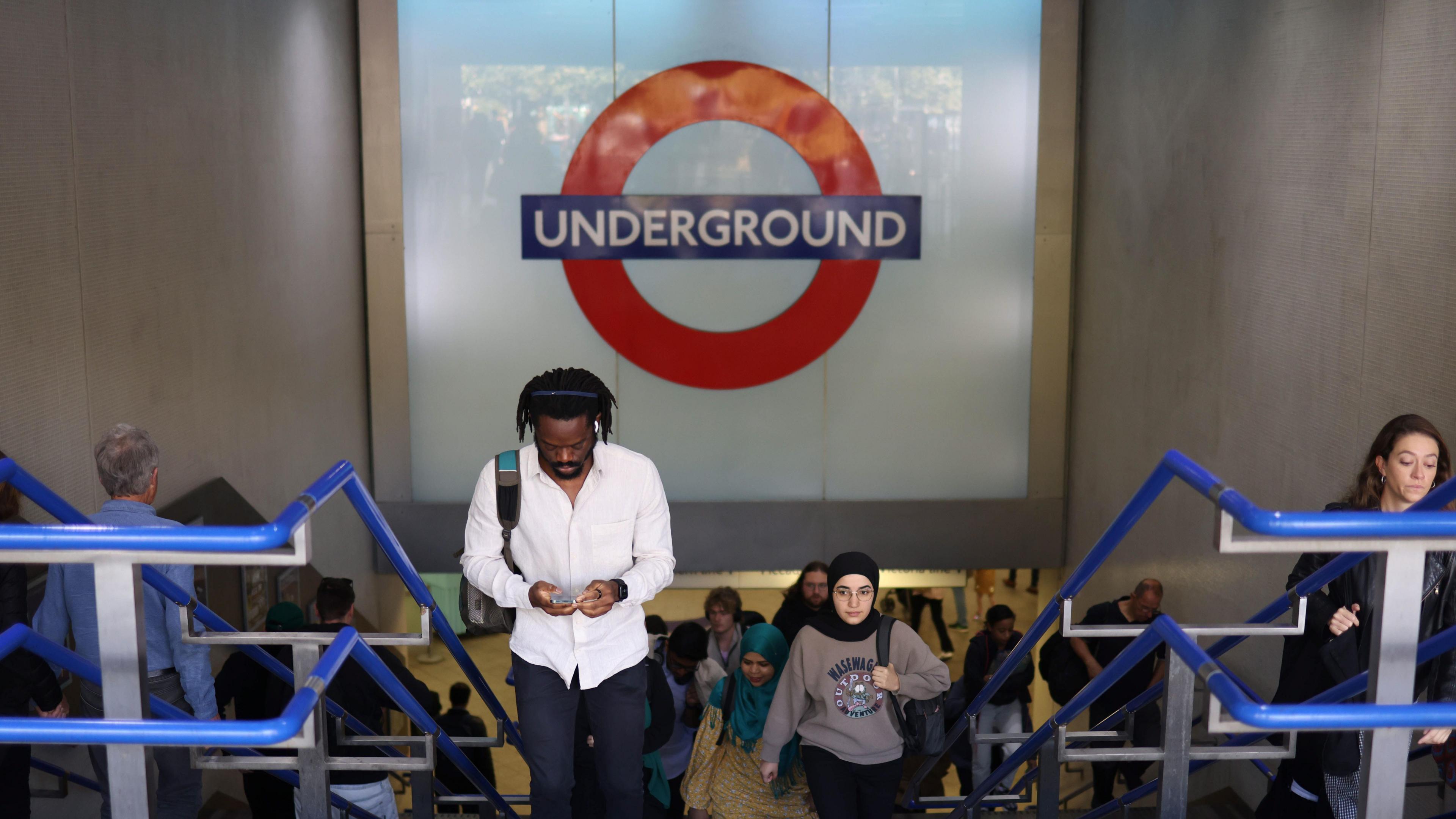 Passengers climbing stairs in a London tube station with a large Underground sign behind them