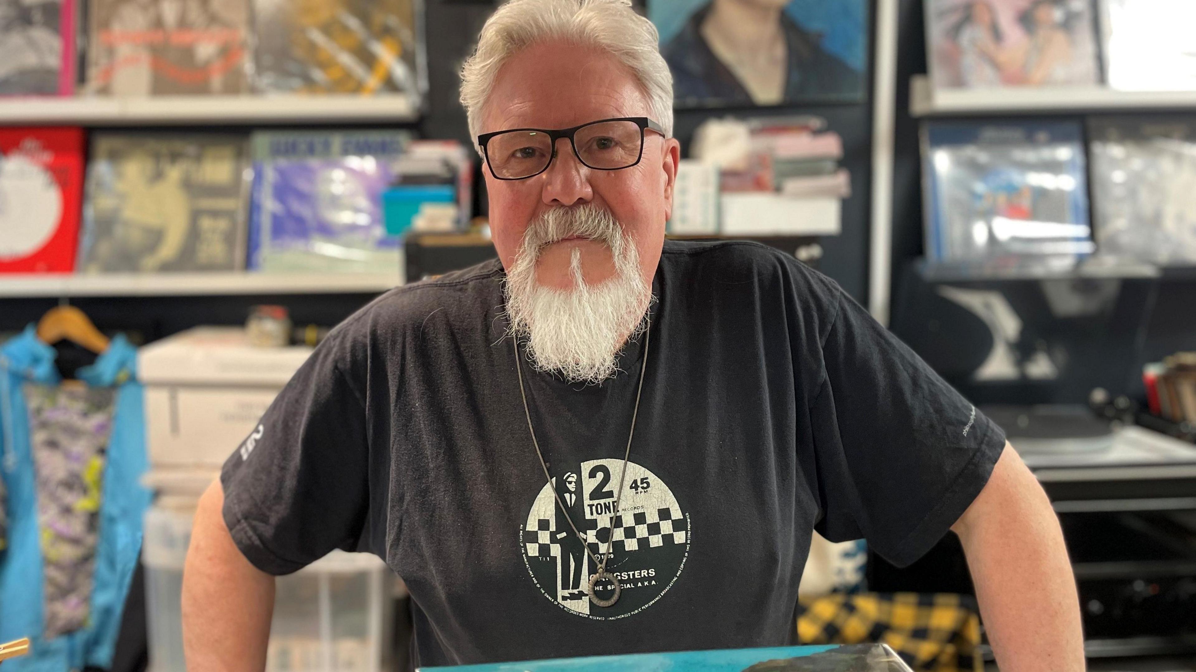 A man with white hair and a beard wearing a band t-shirt stands behind a shop counter