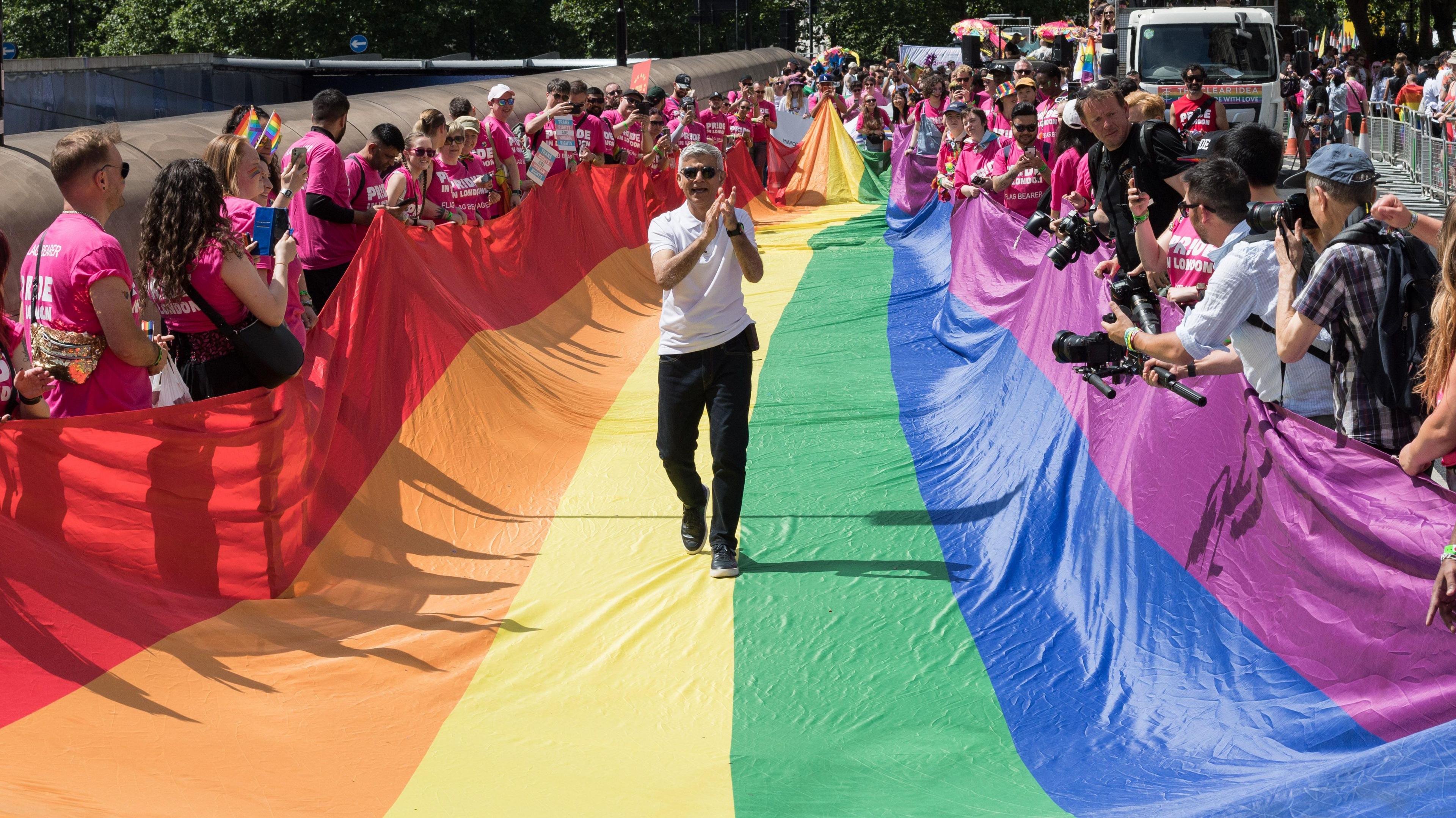 Sadiq Khan, mayor of London walking on top of a large rainbow banner held by people celebrating on both sides. 